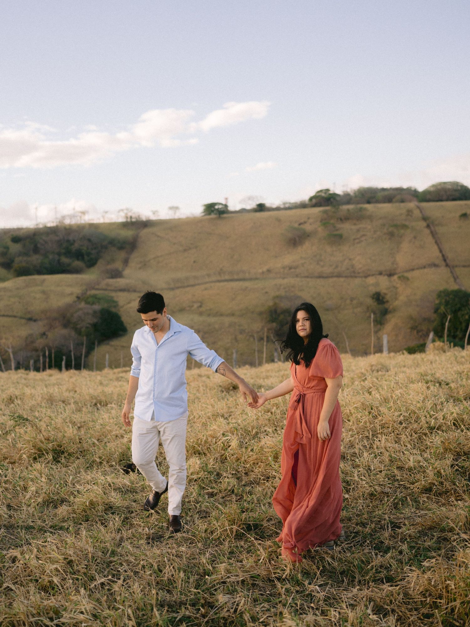 A couple in white shirt and coral dress dance playfully in a grassy hilltop field at sunset.