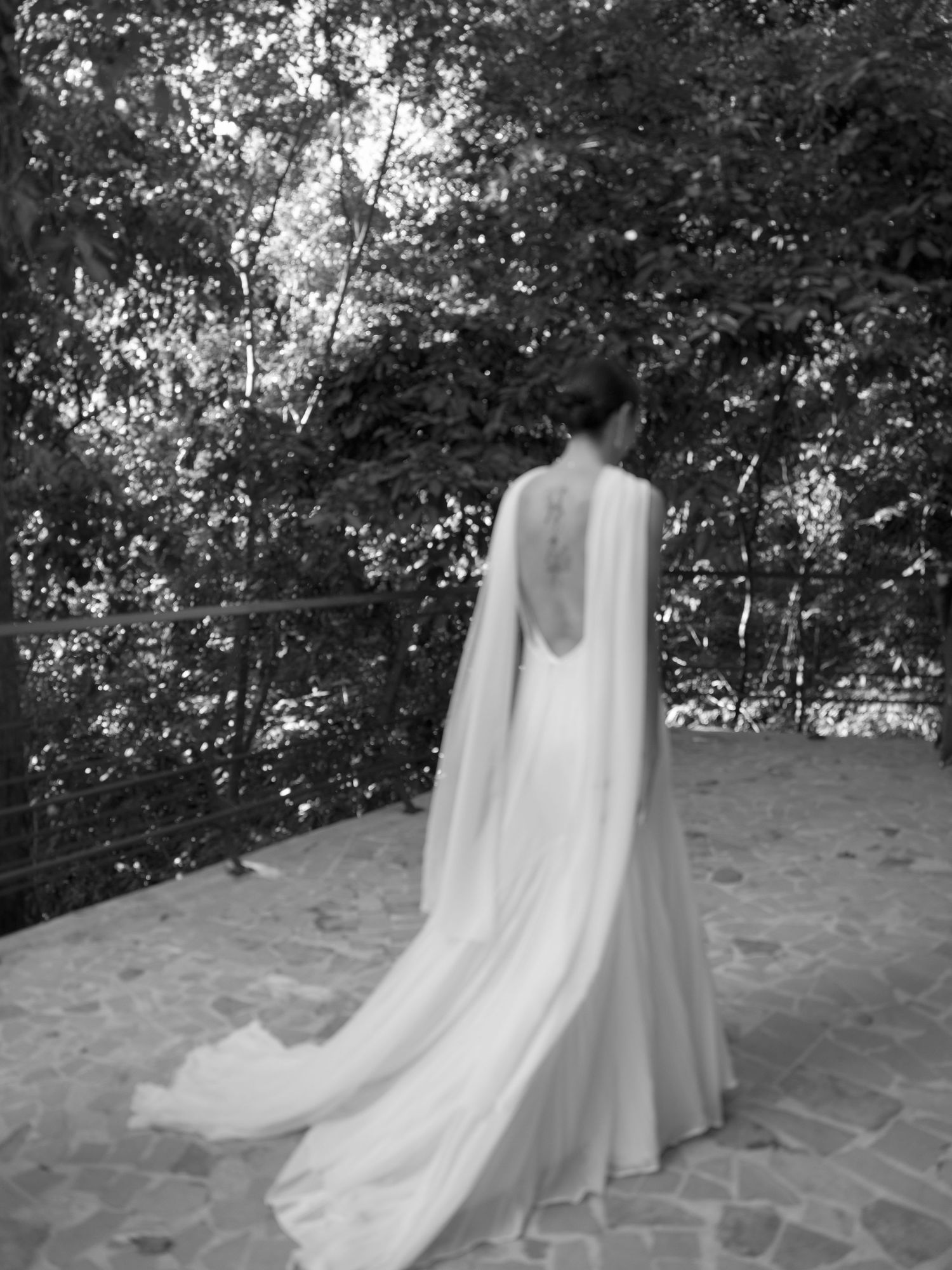 Elegant wedding dress with dramatic open back and flowing cape photographed in black and white against garden backdrop.