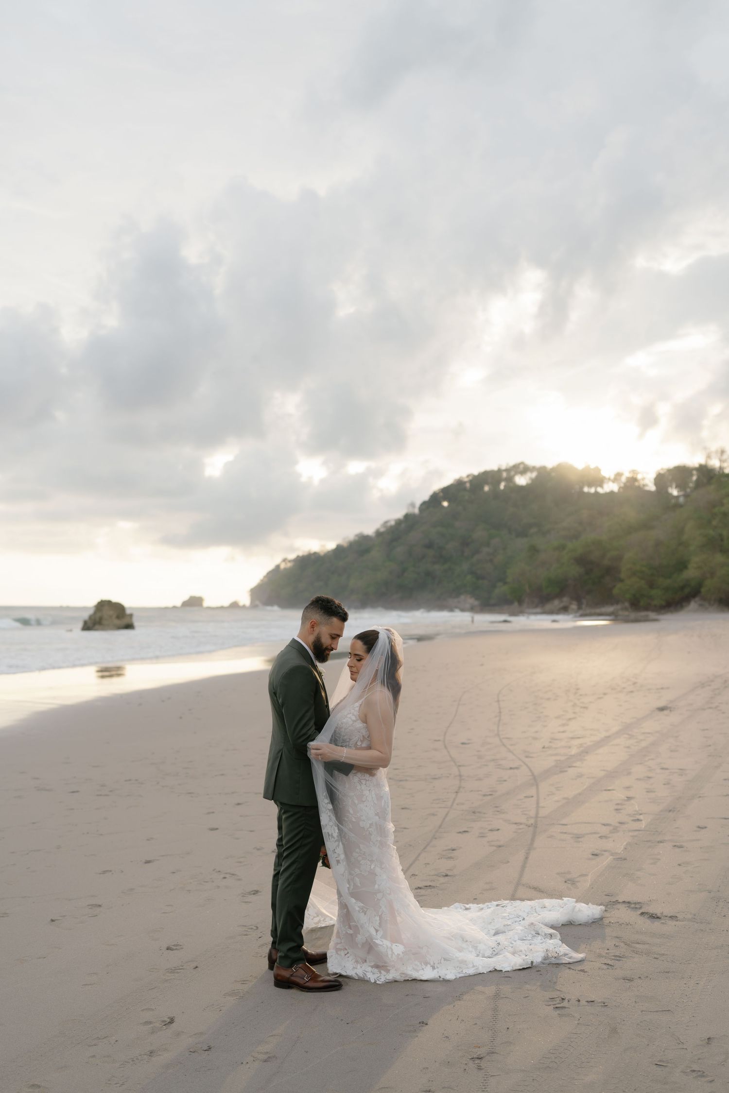 Romantic beach wedding portrait with couple embracing against cloudy sunset sky.