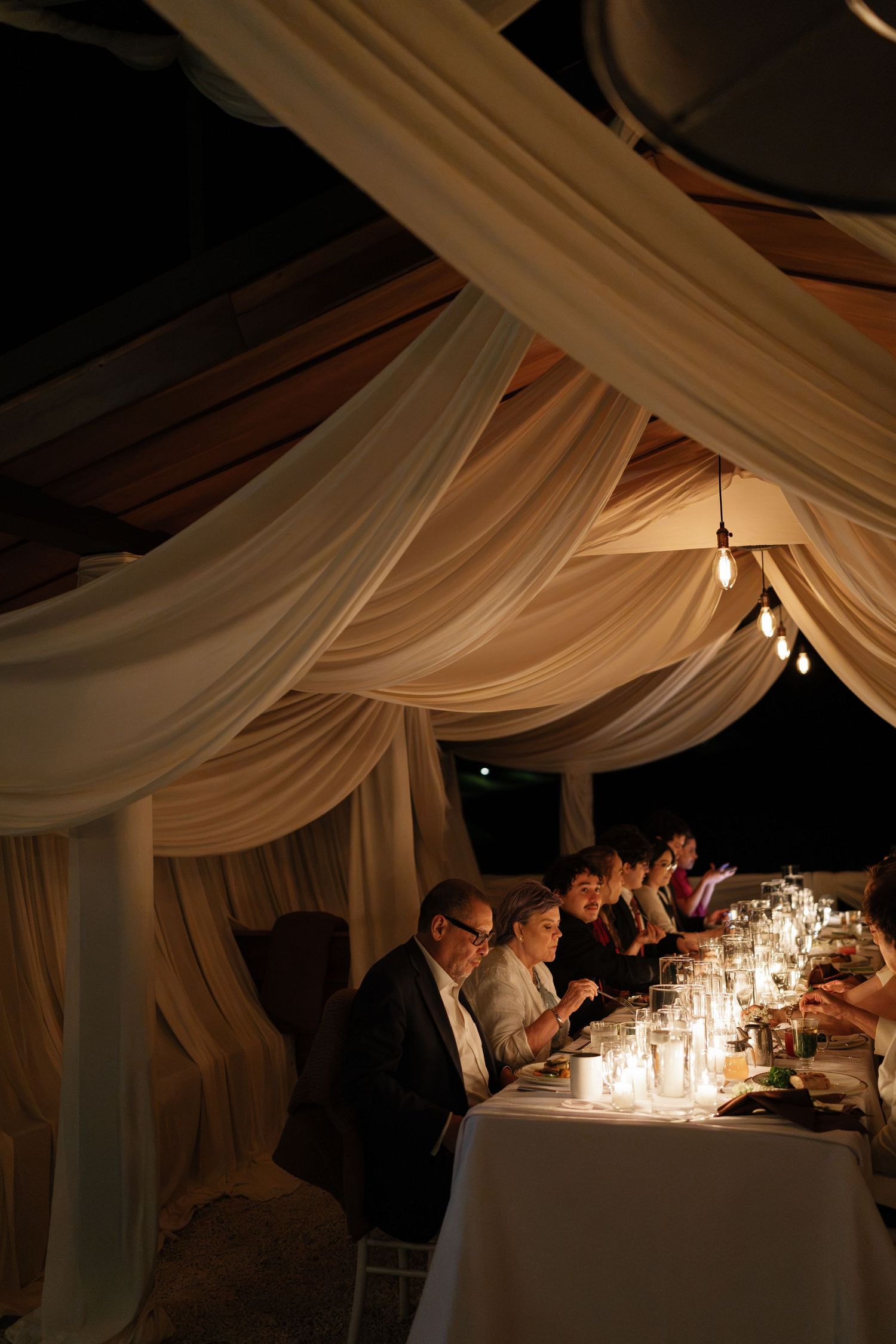 Elegant candlelit dinner party under draped fabric tent with guests seated at long table at night.