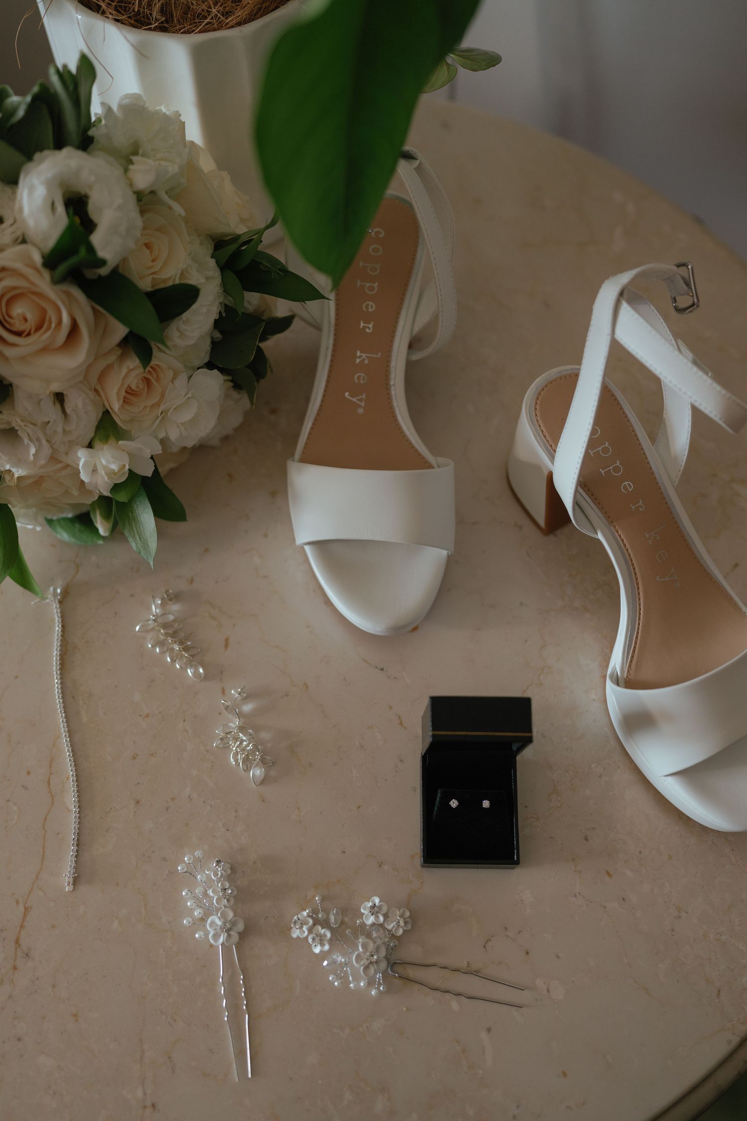 Wedding accessories including white heels, jewelry, and floral arrangements on a beige surface.