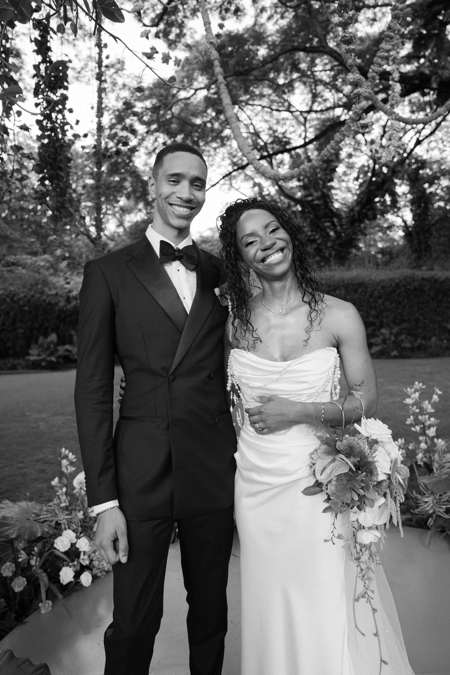 A couple in formal wedding attire smiles together in a garden setting.