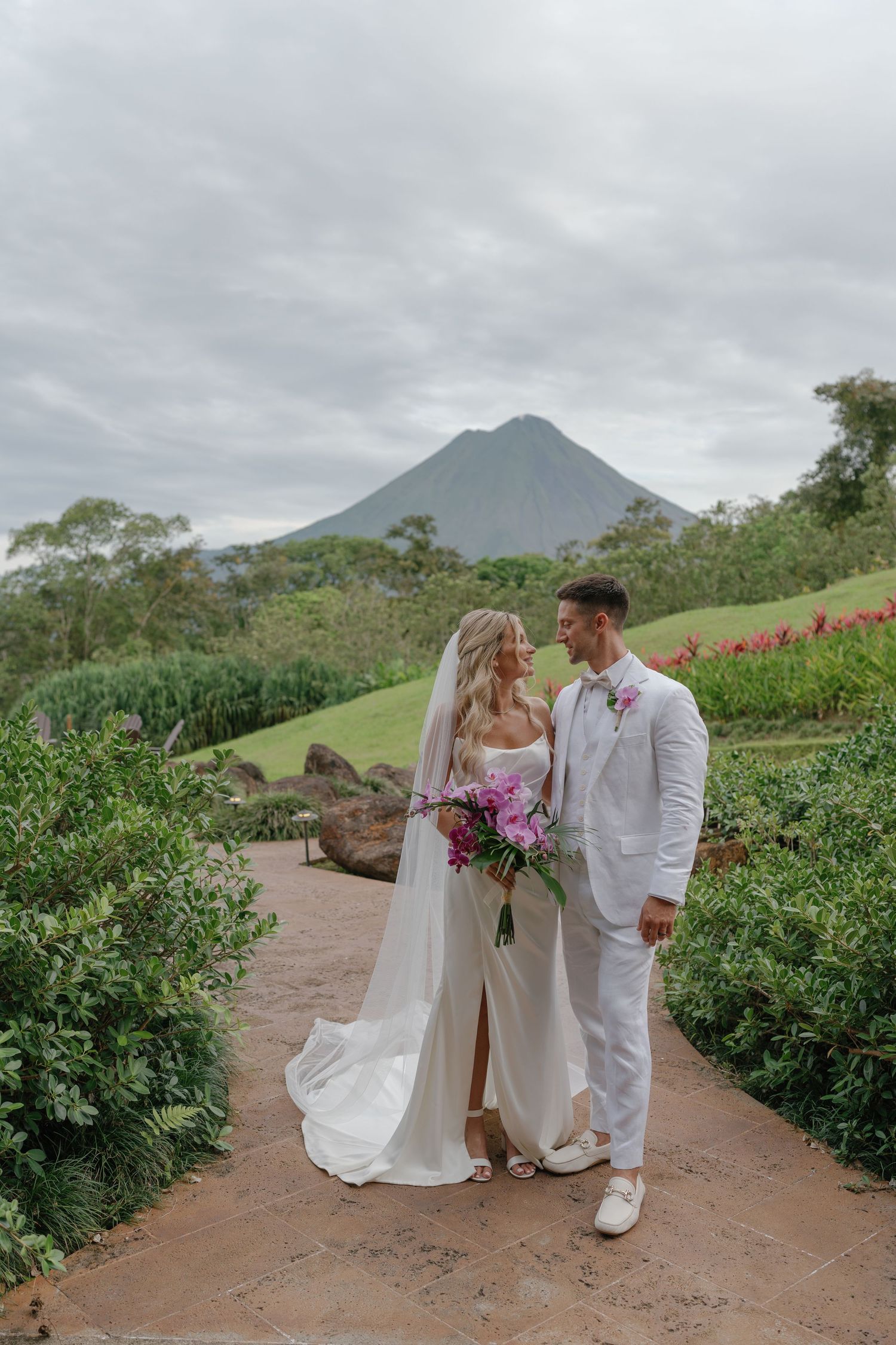 Couple walks along mountain path with volcanic peak visible in background.