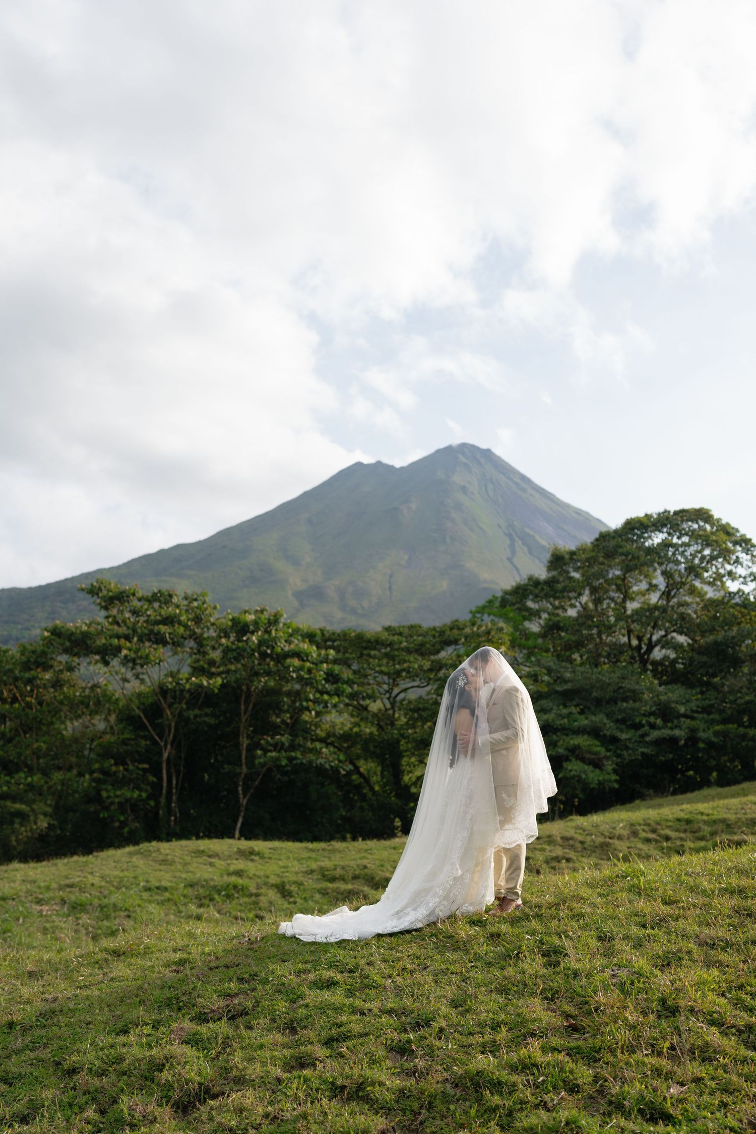 Wedding couple photographed from afar on green hillside with majestic volcanic peaks.