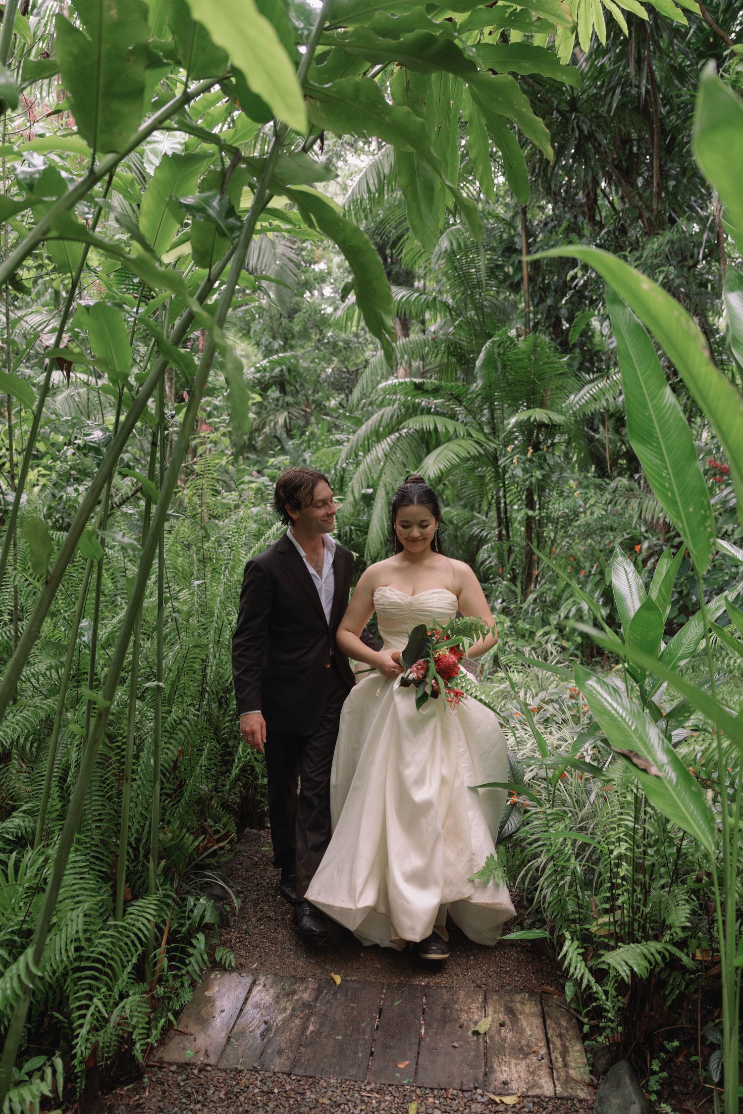 A couple in wedding attire walks along a wooden path surrounded by tropical foliage.