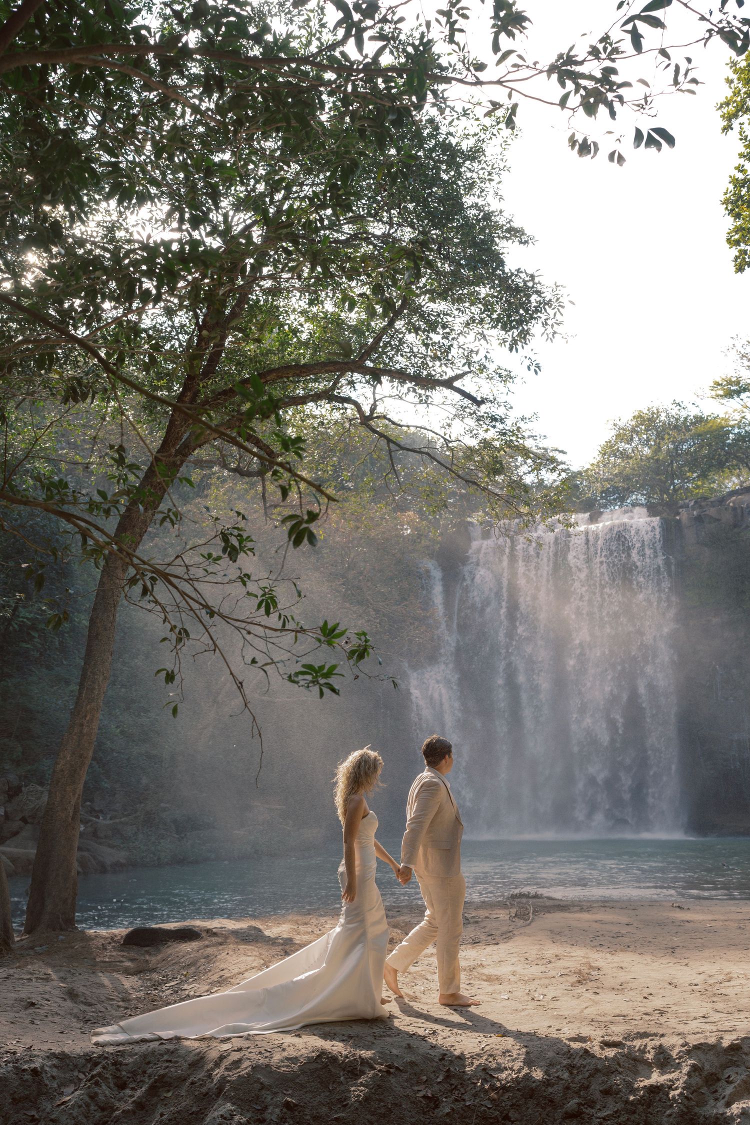 Two ethereal figures in white dresses stand near a misty waterfall in a dreamy forest setting.
