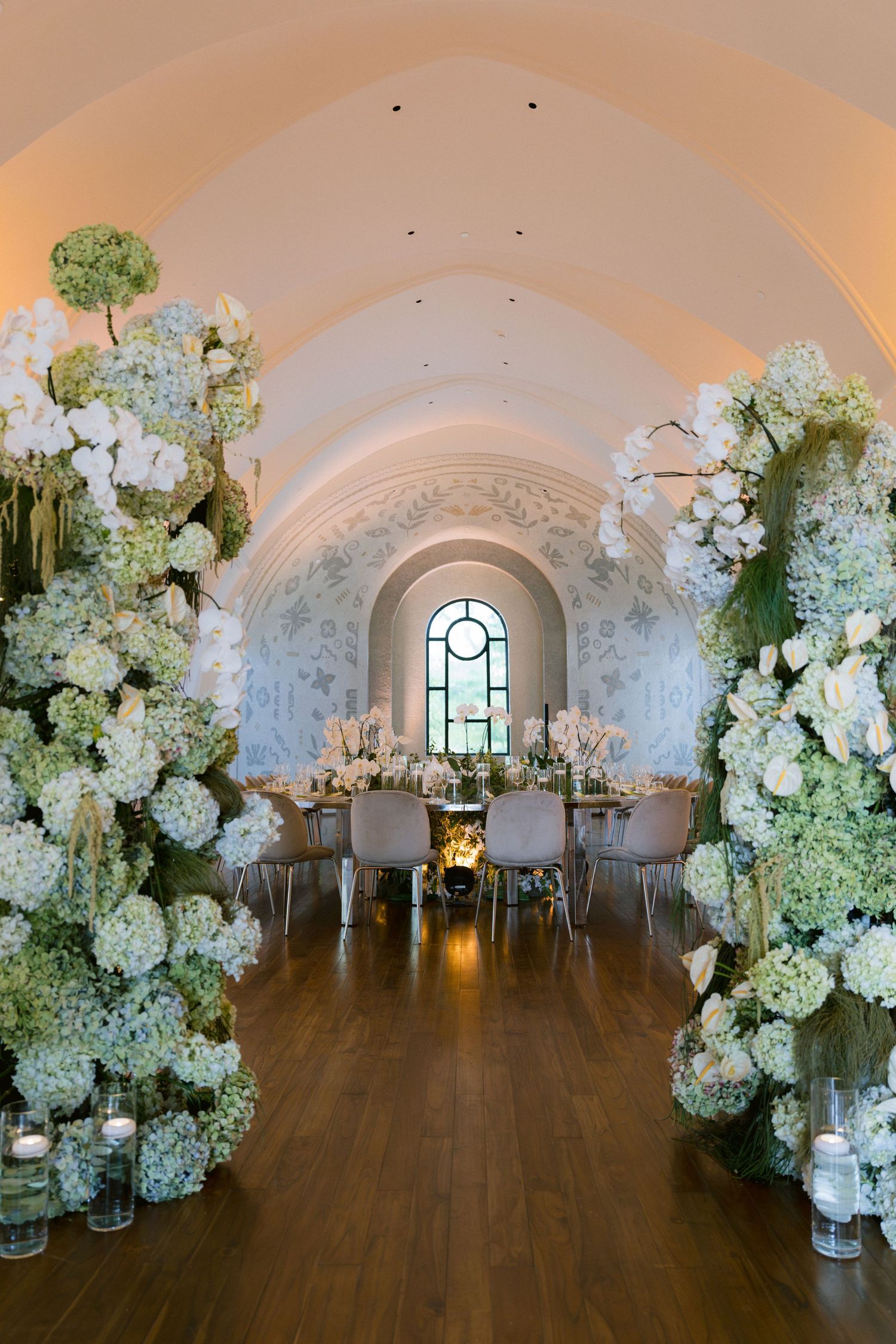 Elegant wedding reception hall with lush white hydrangea floral arches, long dining table, candlelight, and arched ceiling.