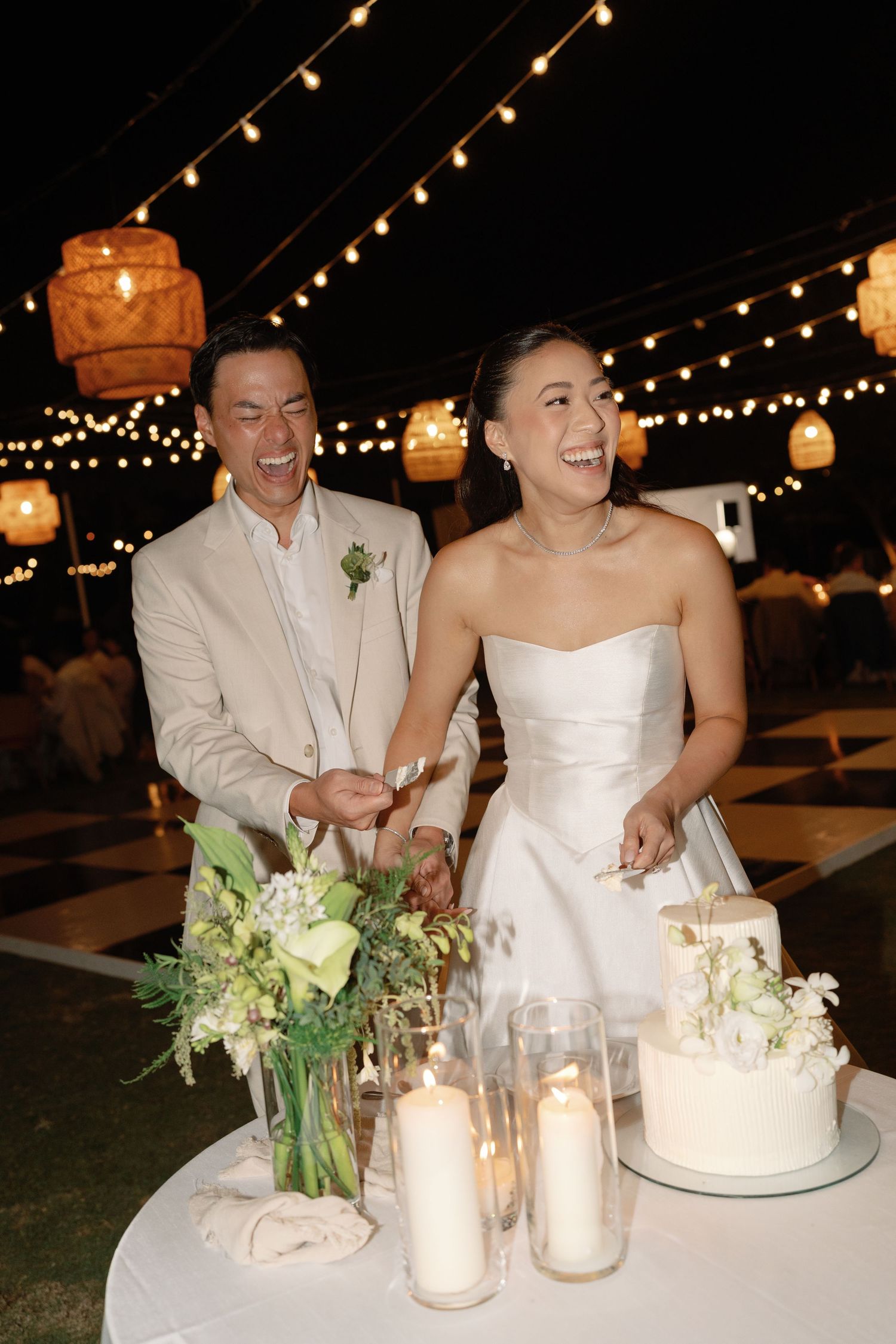 A couple cutting their wedding cake together under string lights at their outdoor reception.