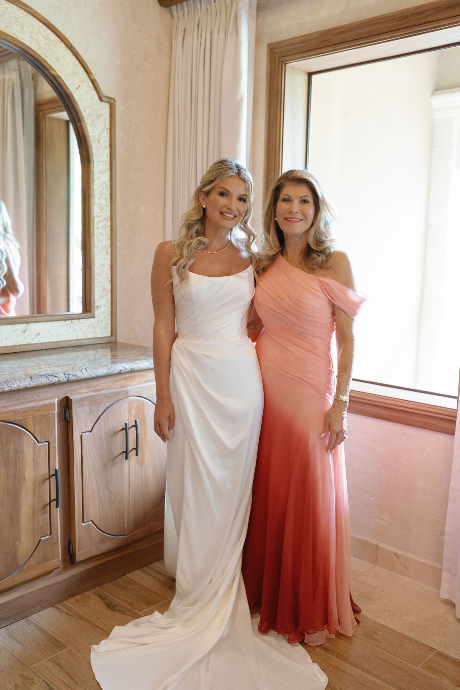 Two people pose together in white and coral dresses in an elegant room with wood floors.