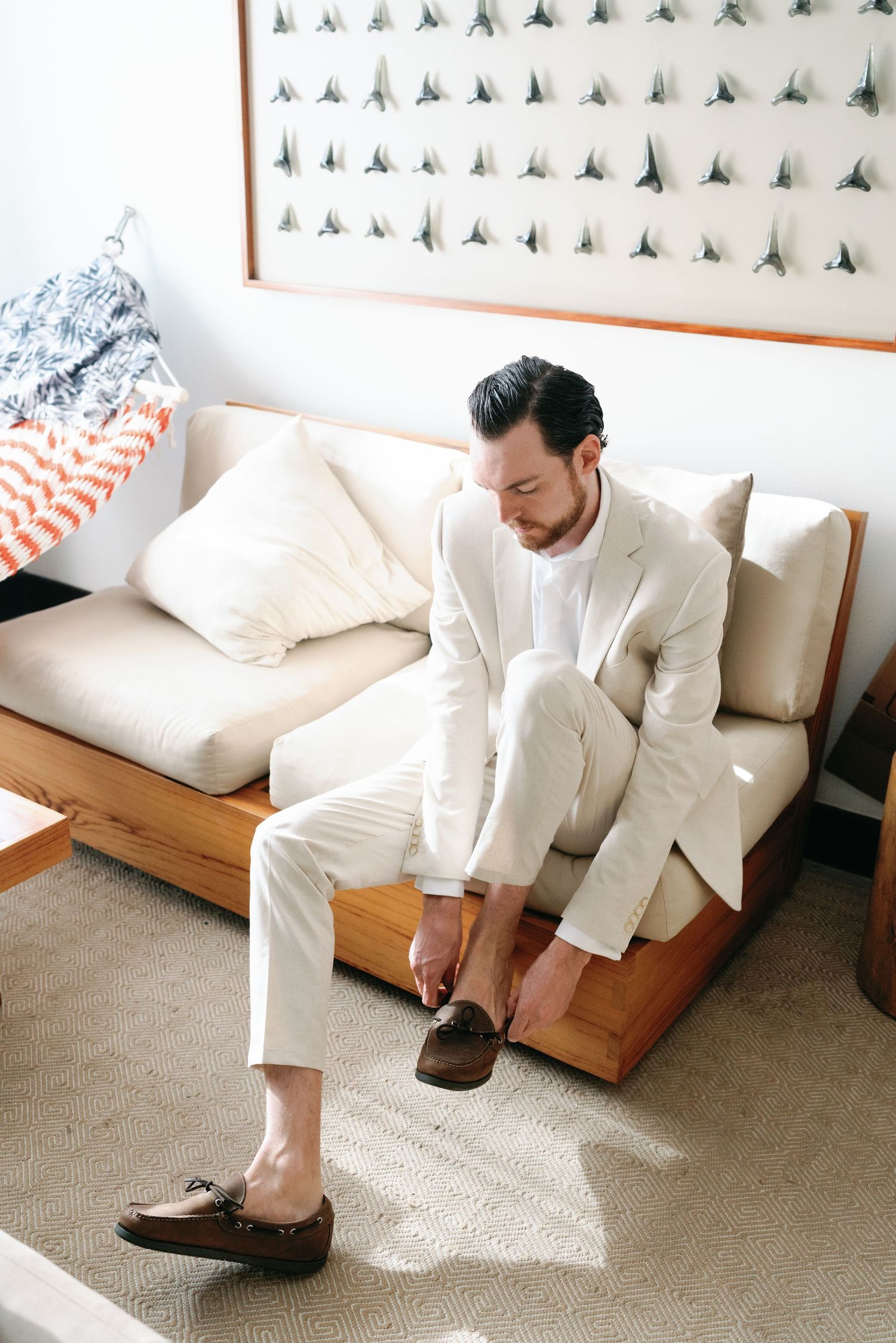 Close-up shots of groom putting on brown dress shoes while wearing a white suit on a beige couch.