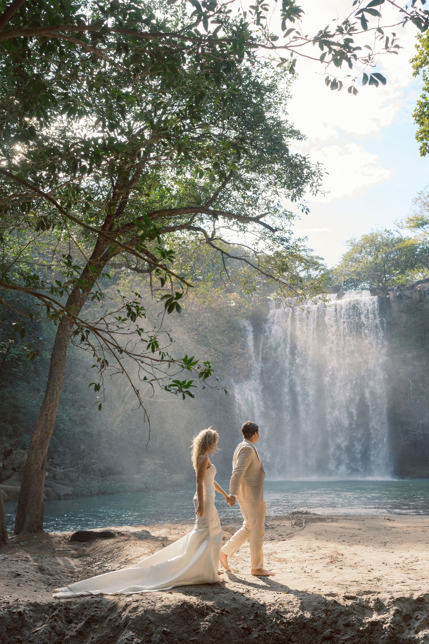 A couple in white attire poses gracefully by a majestic waterfall in a dreamy natural setting.