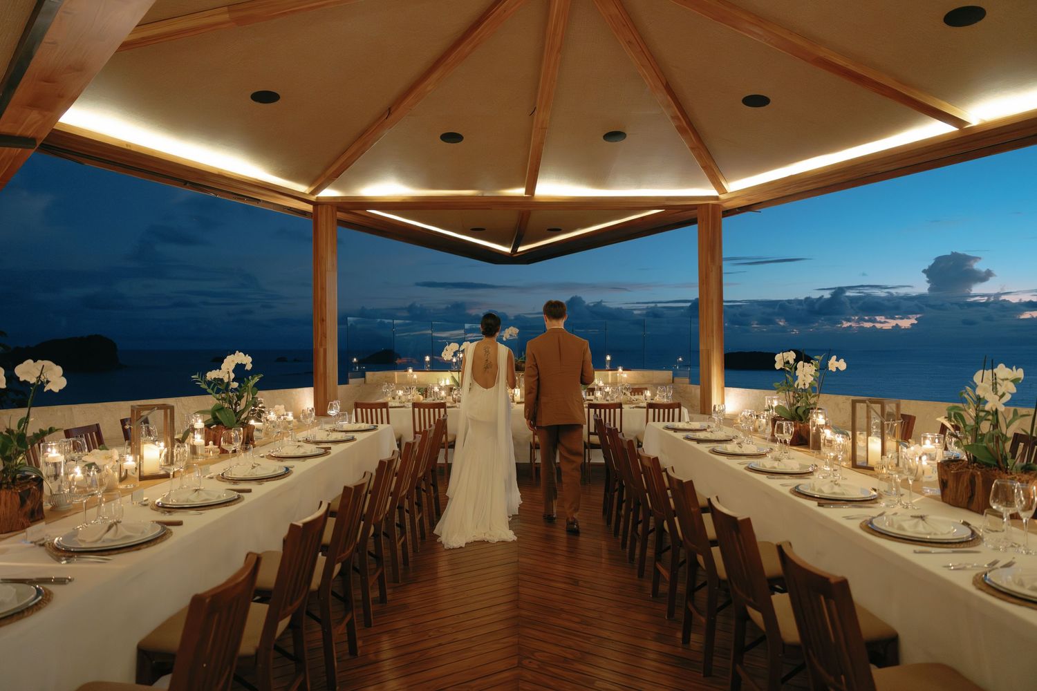 Elegant outdoor wedding reception setup under wooden pavilion at sunset with ocean views and long dining tables.