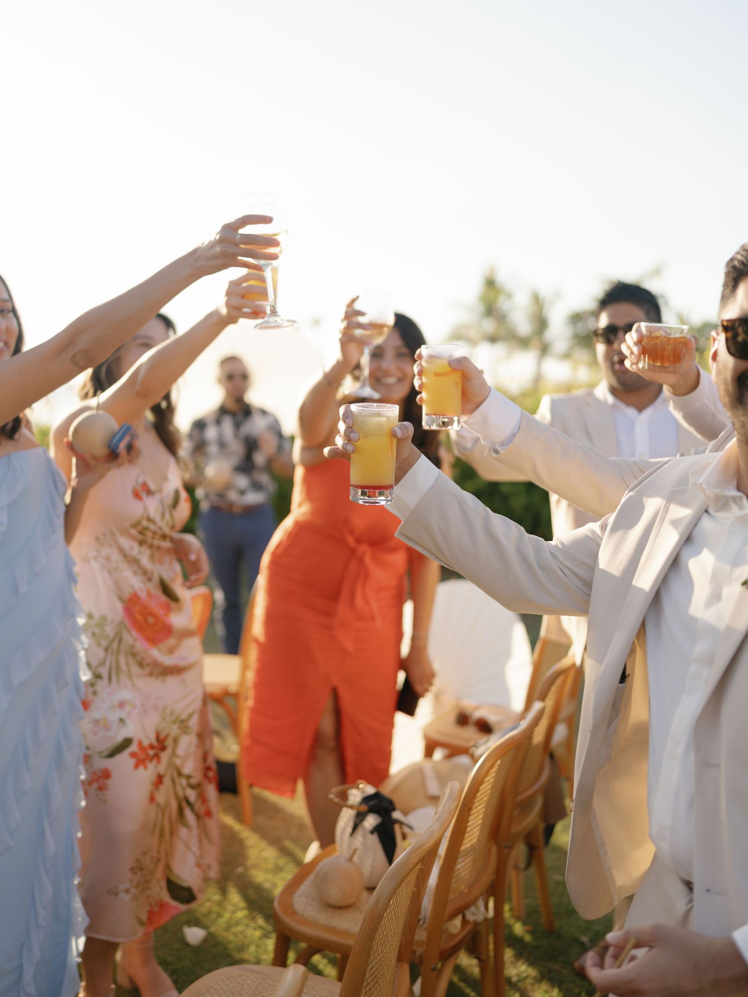 Wedding guests in white and orange attire raising glasses for a celebratory toast at sunset.