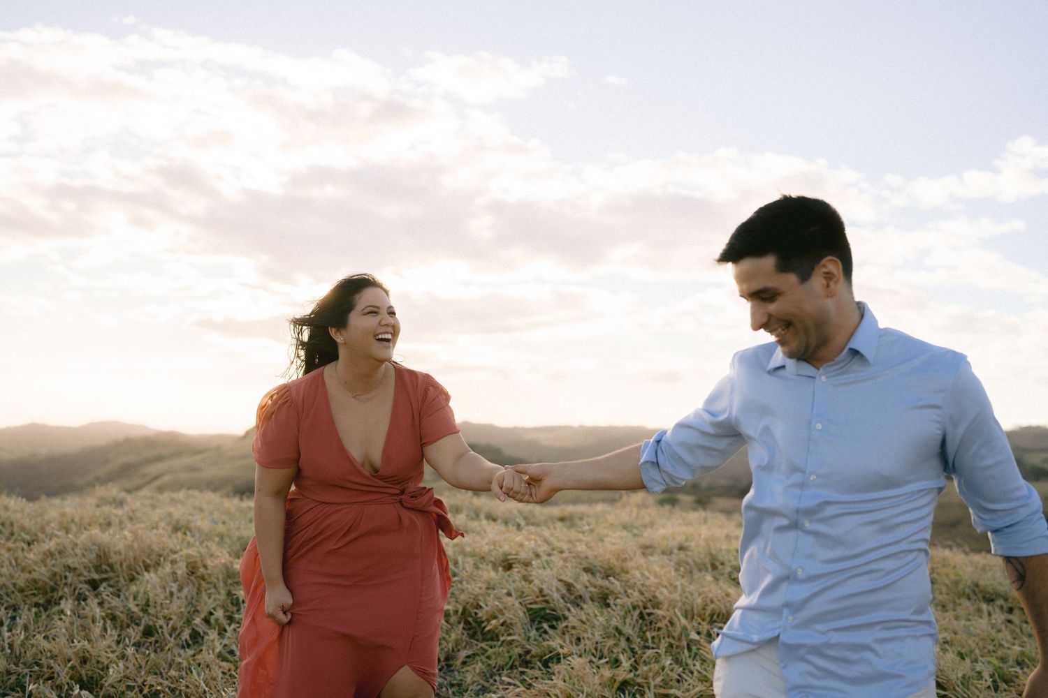 A couple embraces during sunset on a grassy hilltop with mountains in the background.