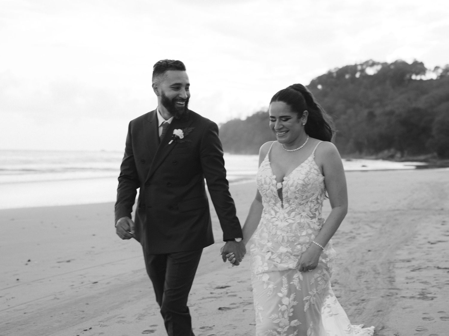 A romantic black and white photo of a couple walking along a scenic beach in formal attire.