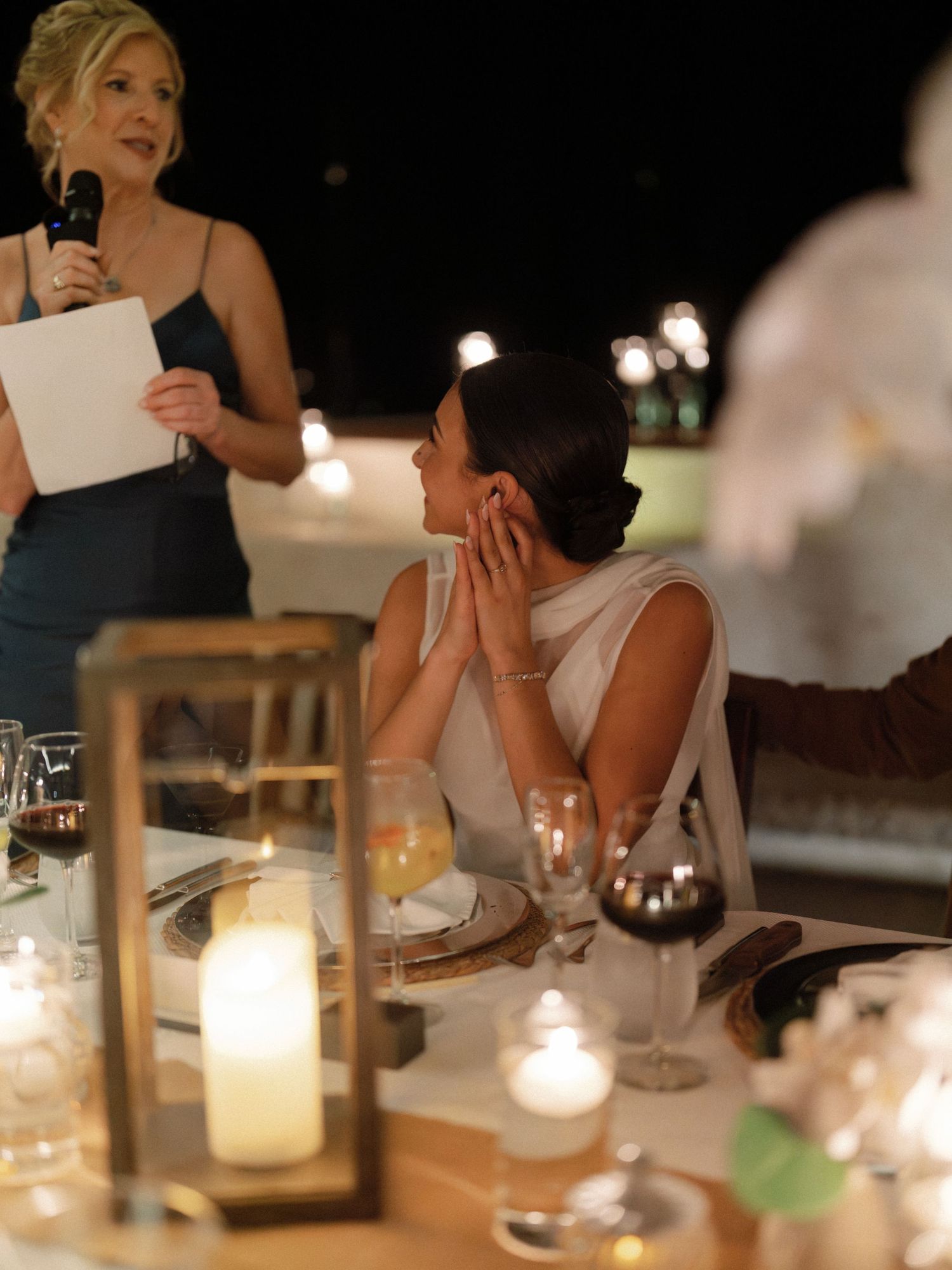 A candlelit wedding reception table scene with someone giving a speech while another person reacts emotionally.