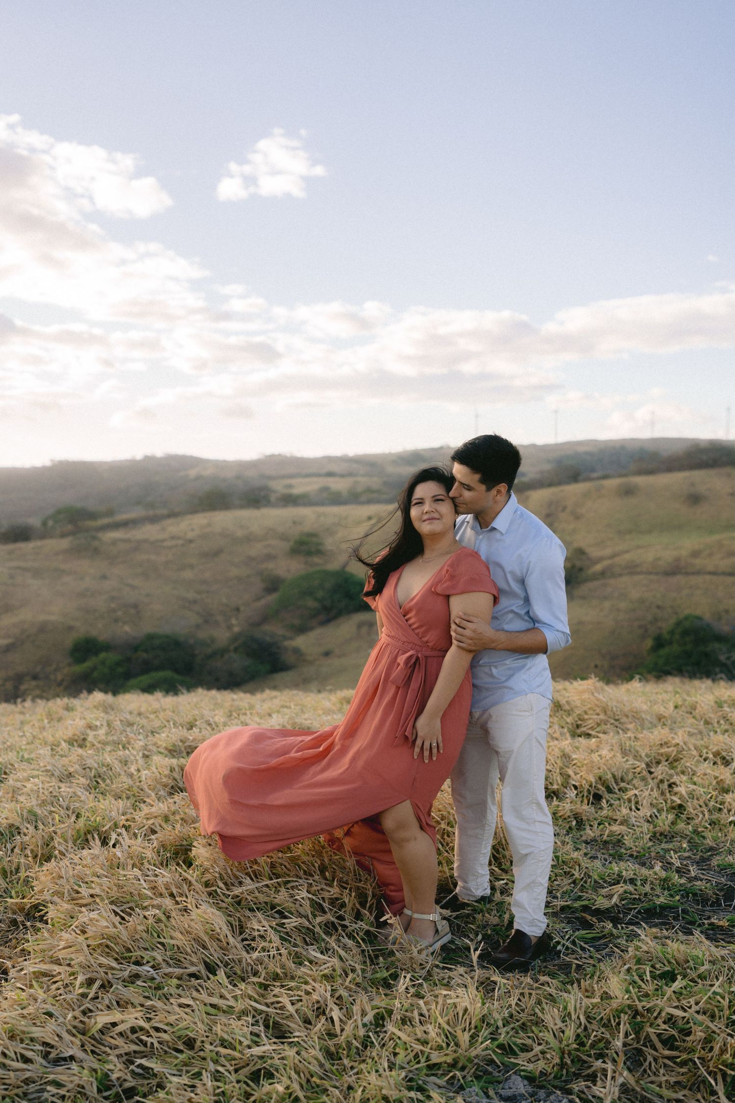 A flowing coral dress catches the wind as a couple embraces on a hilltop during golden hour.