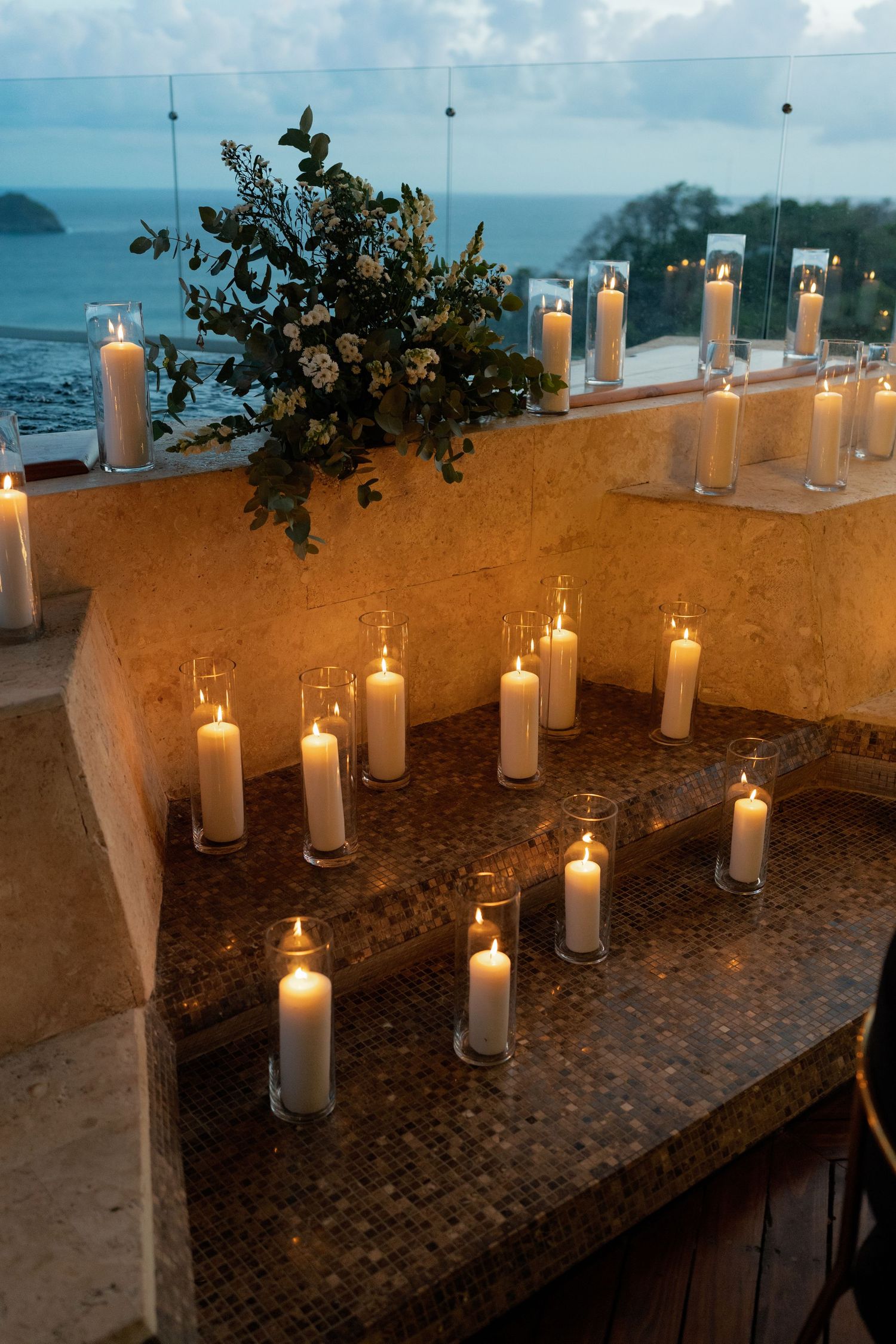 White pillar candles arranged on a stone wall overlooking an ocean view at dusk.