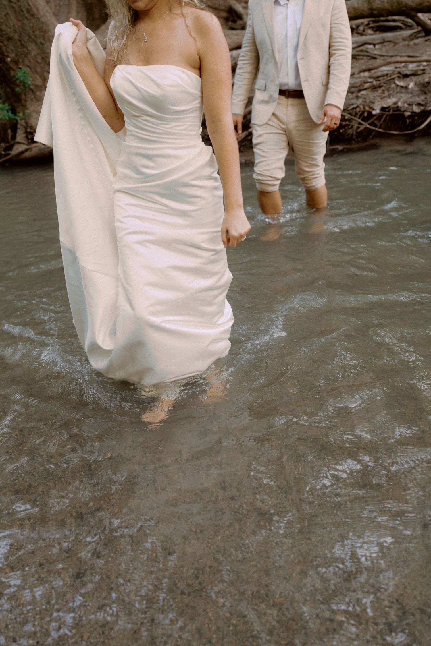 Two people in white clothing wade through shallow water near a rocky shore.
