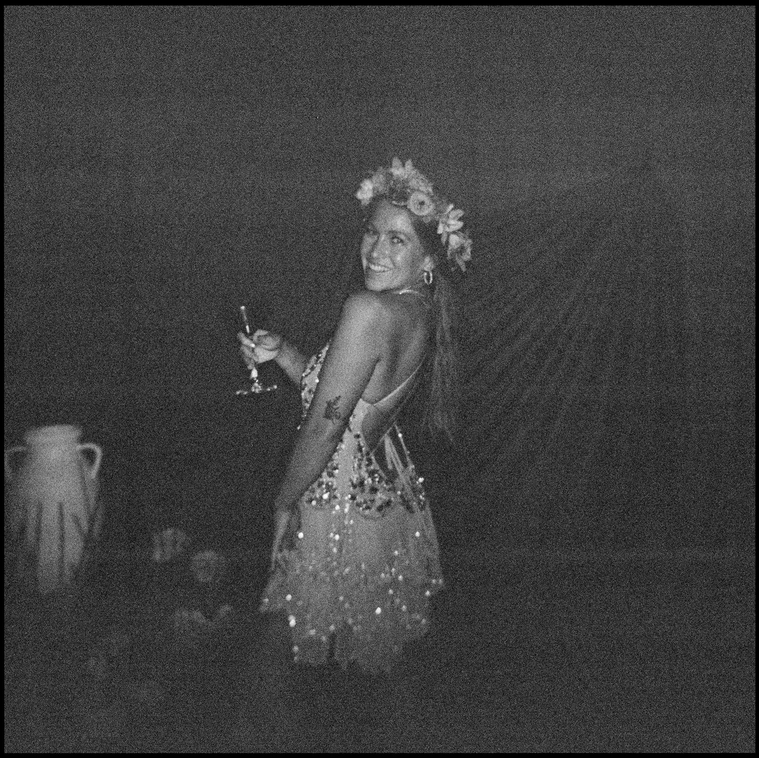 Black and white photo of a smiling woman in a floral crown and sparkly dress holding a cocktail glass at a party.
