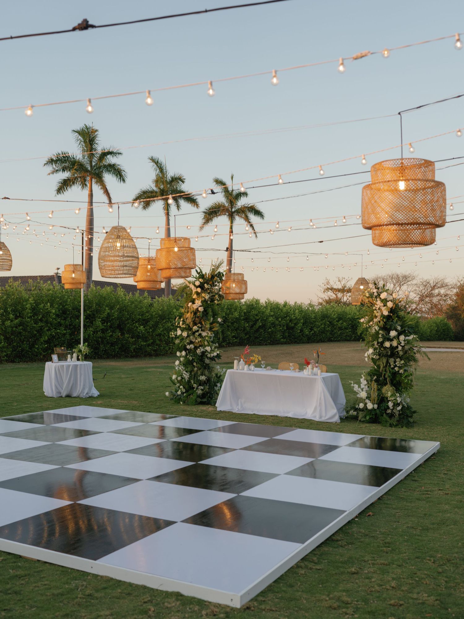 An outdoor wedding reception setup with checkered dance floor and hanging rattan lanterns at dusk.