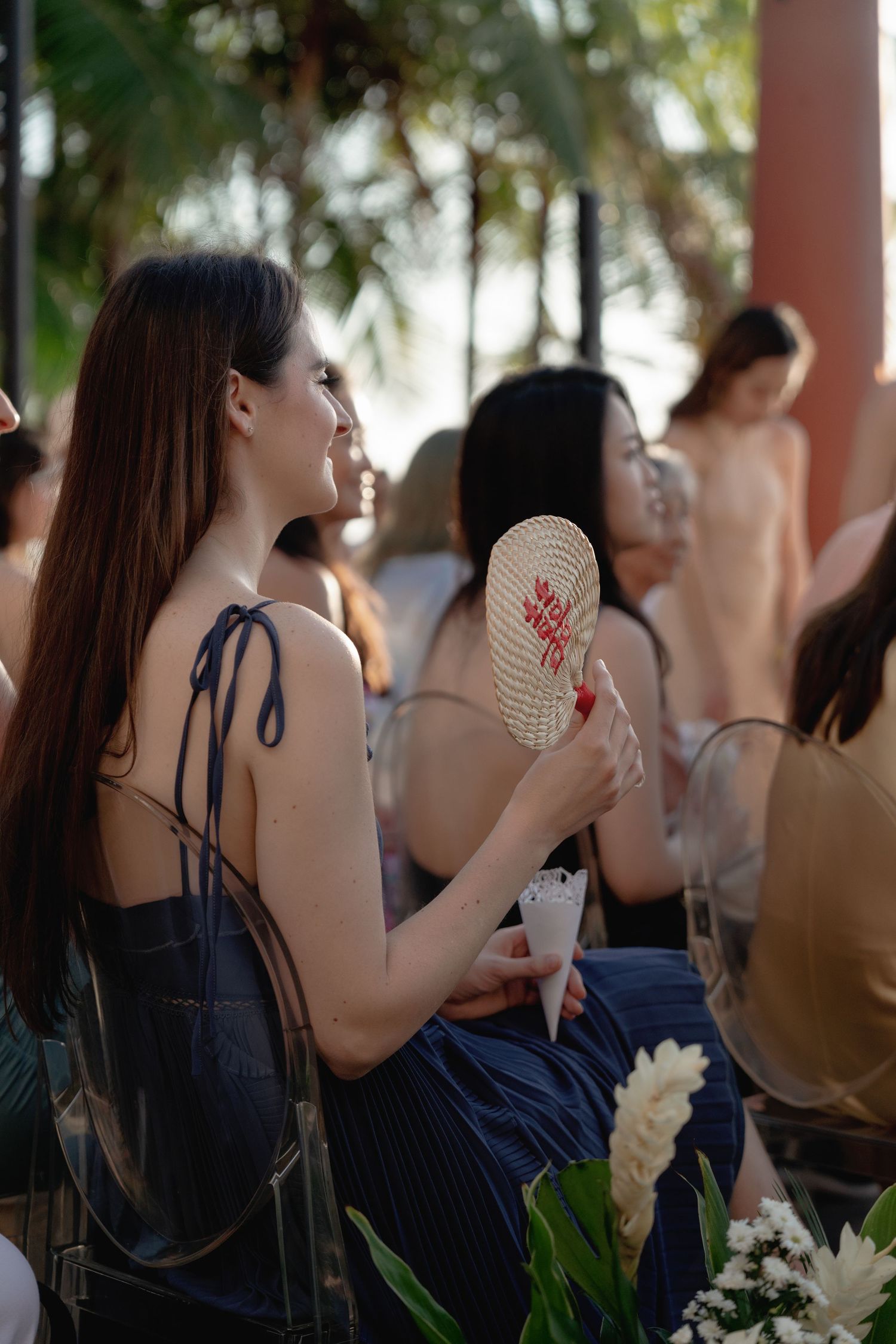 A group of wedding guests seated at an outdoor reception venue with palm trees and floral decorations visible.
