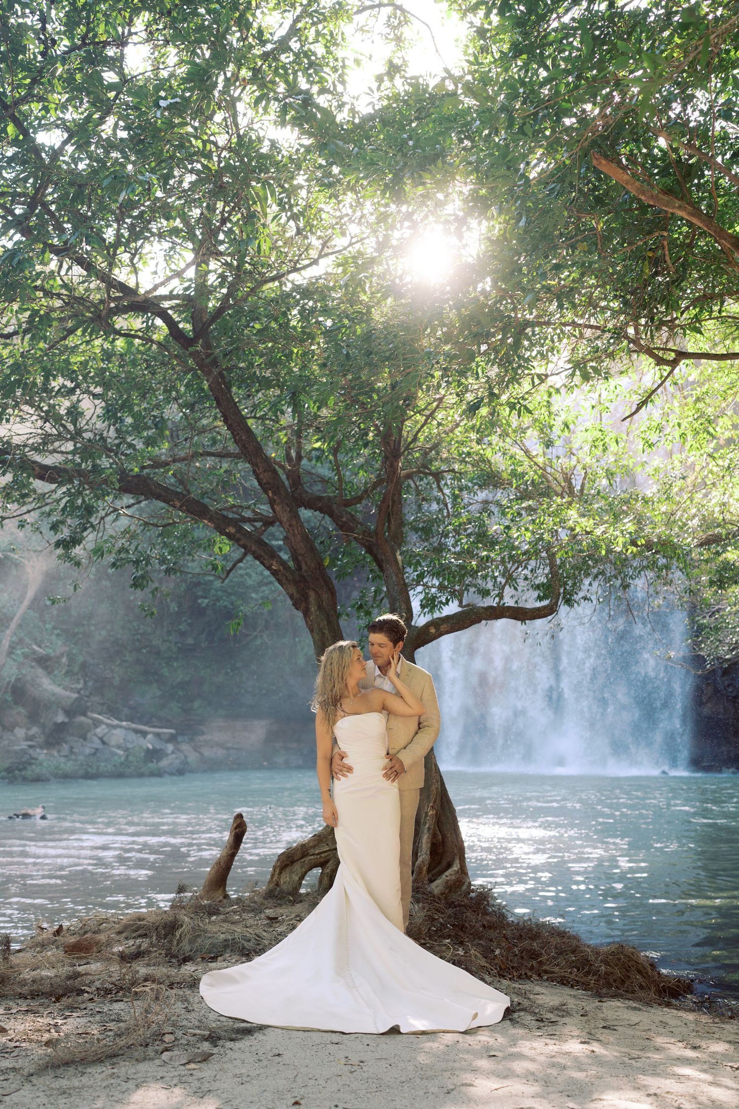 A romantic wedding photoshoot by a scenic waterfall with sunlight filtering through trees.