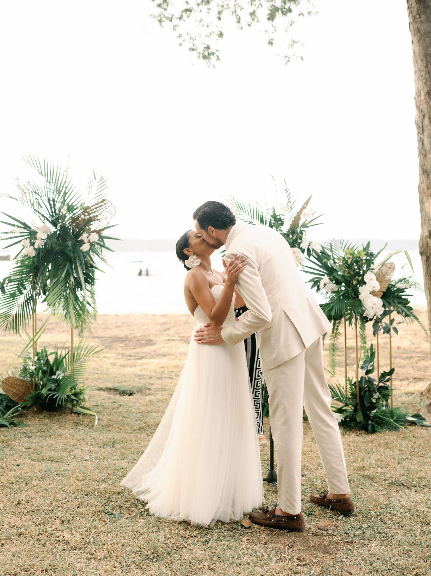A romantic beach wedding photoshoot featuring a couple in white attire posing among tropical palm plants.