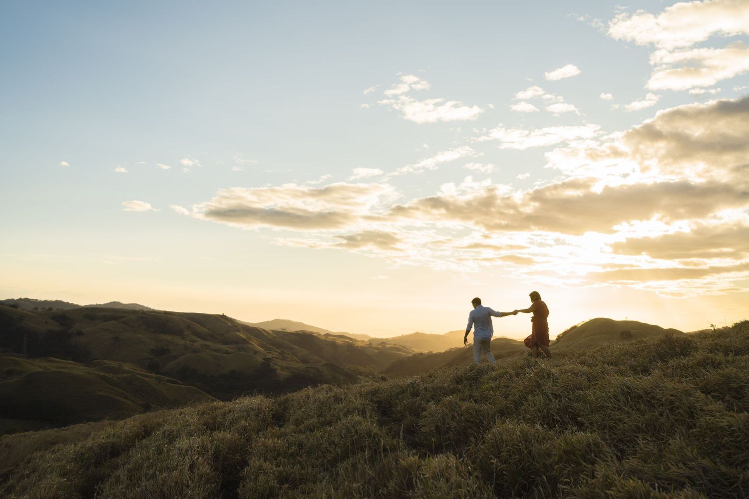 Silhouettes hold hands on a hilltop during a dramatic sunset with mountains in the distance.