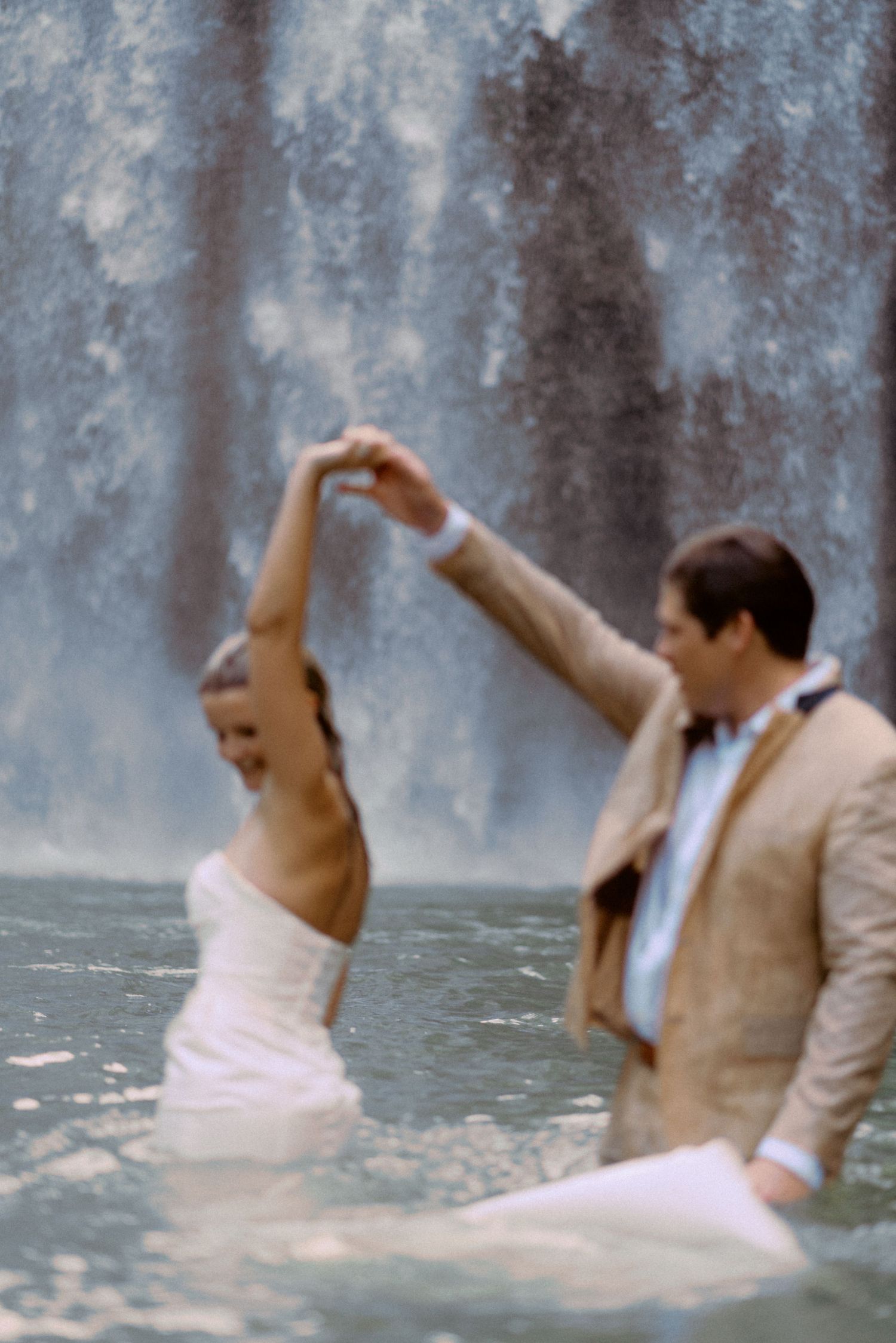 A couple playfully falls backwards into a waterfall during a romantic photoshoot.