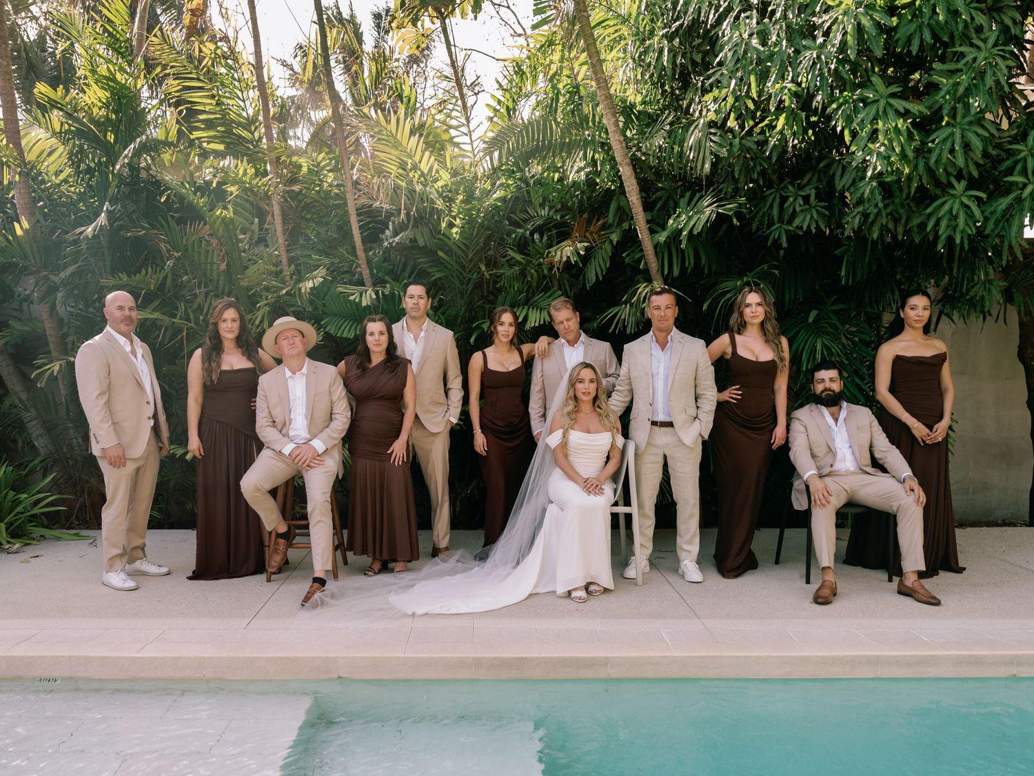 Wedding party members sit casually on stone steps in a tropical garden setting during an outdoor celebration.