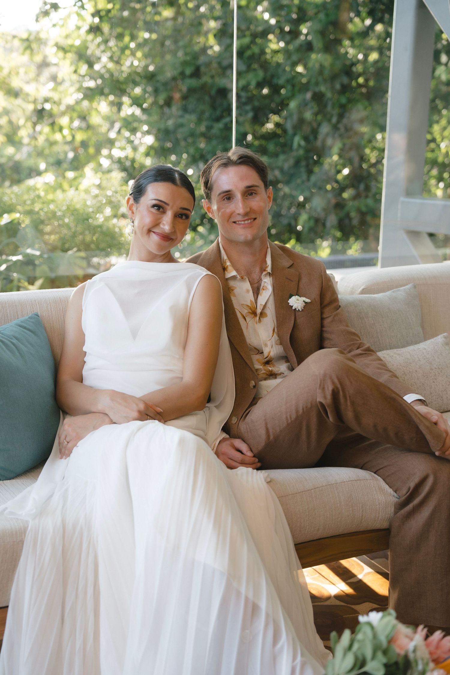 A couple relaxes together on a porch sofa, with the person in white wearing a flowing dress and their companion in brown.