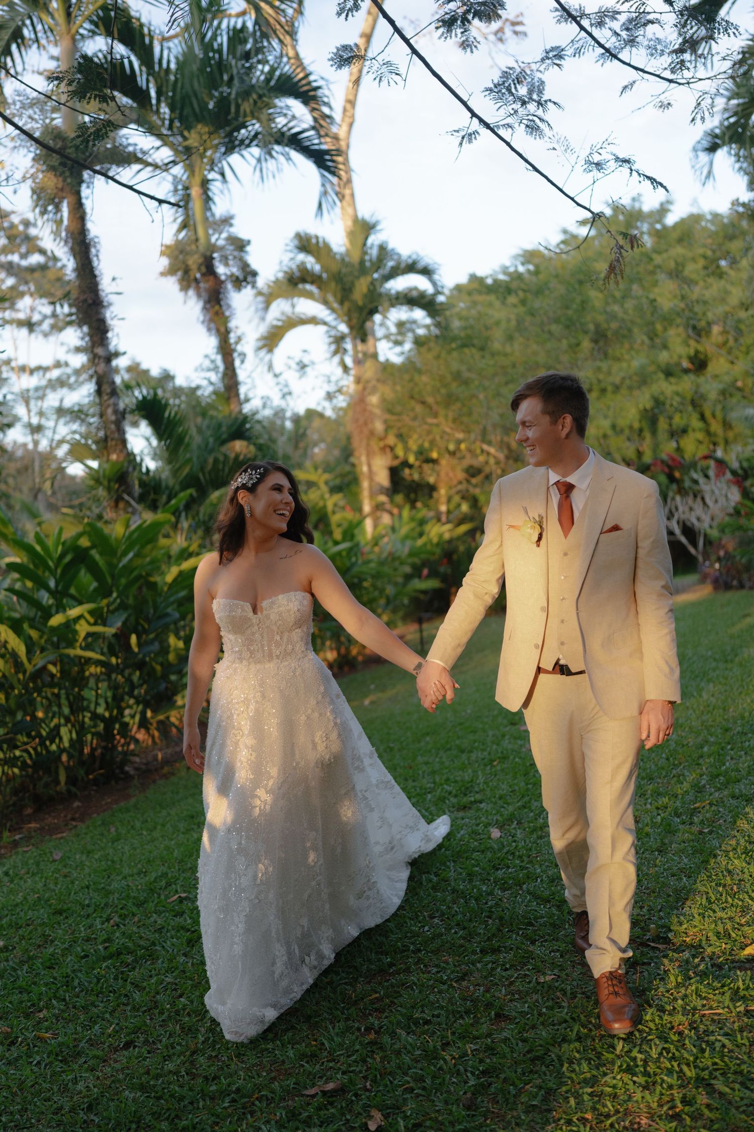 A couple in wedding attire walks through a lush garden path during golden hour.