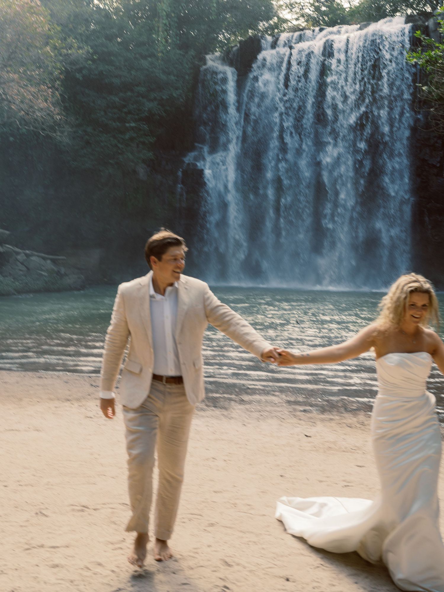 Two people in white formal wear dance joyfully by a tropical waterfall.