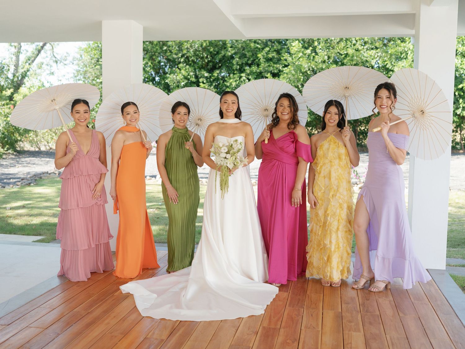 Bridesmaids in colorful pastel dresses line up outdoors under a covered patio area.