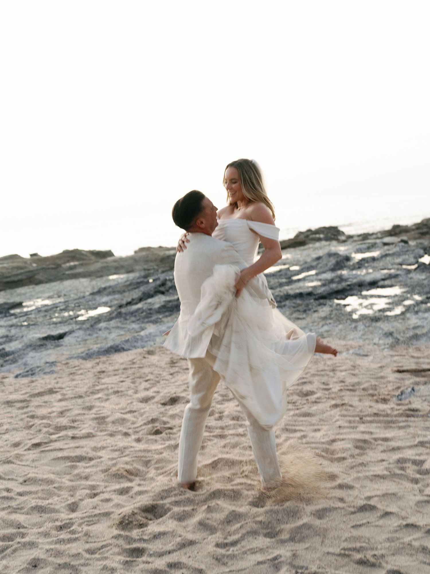 A couple in white attire embrace joyfully on a sandy beach with rocky cliffs in the background during sunset.