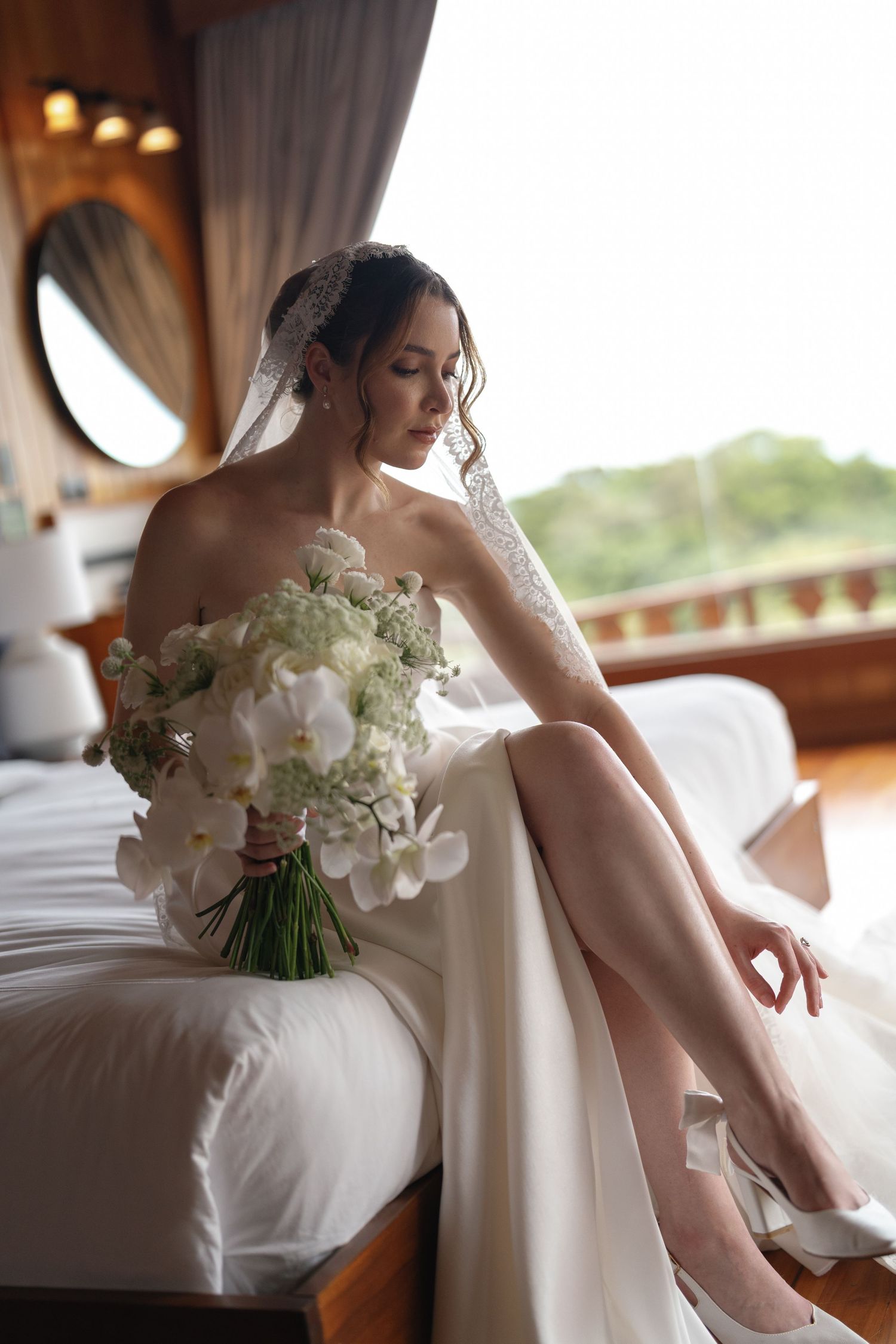 Elegant bride in white strapless gown and veil sits on bed holding white orchid bouquet in a luxury room.