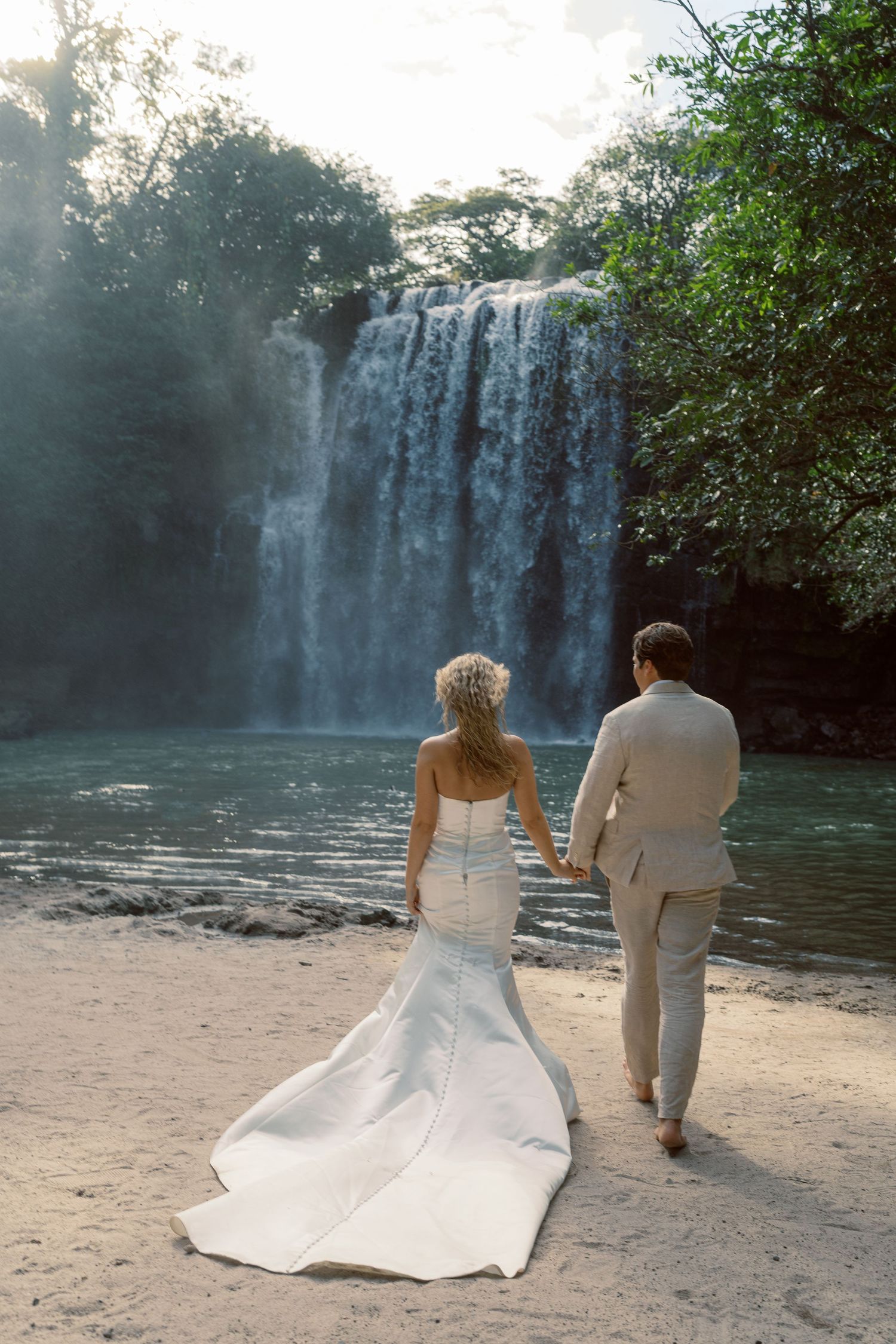 Two figures in formal white attire walk along a sandy shore with a waterfall in the background.