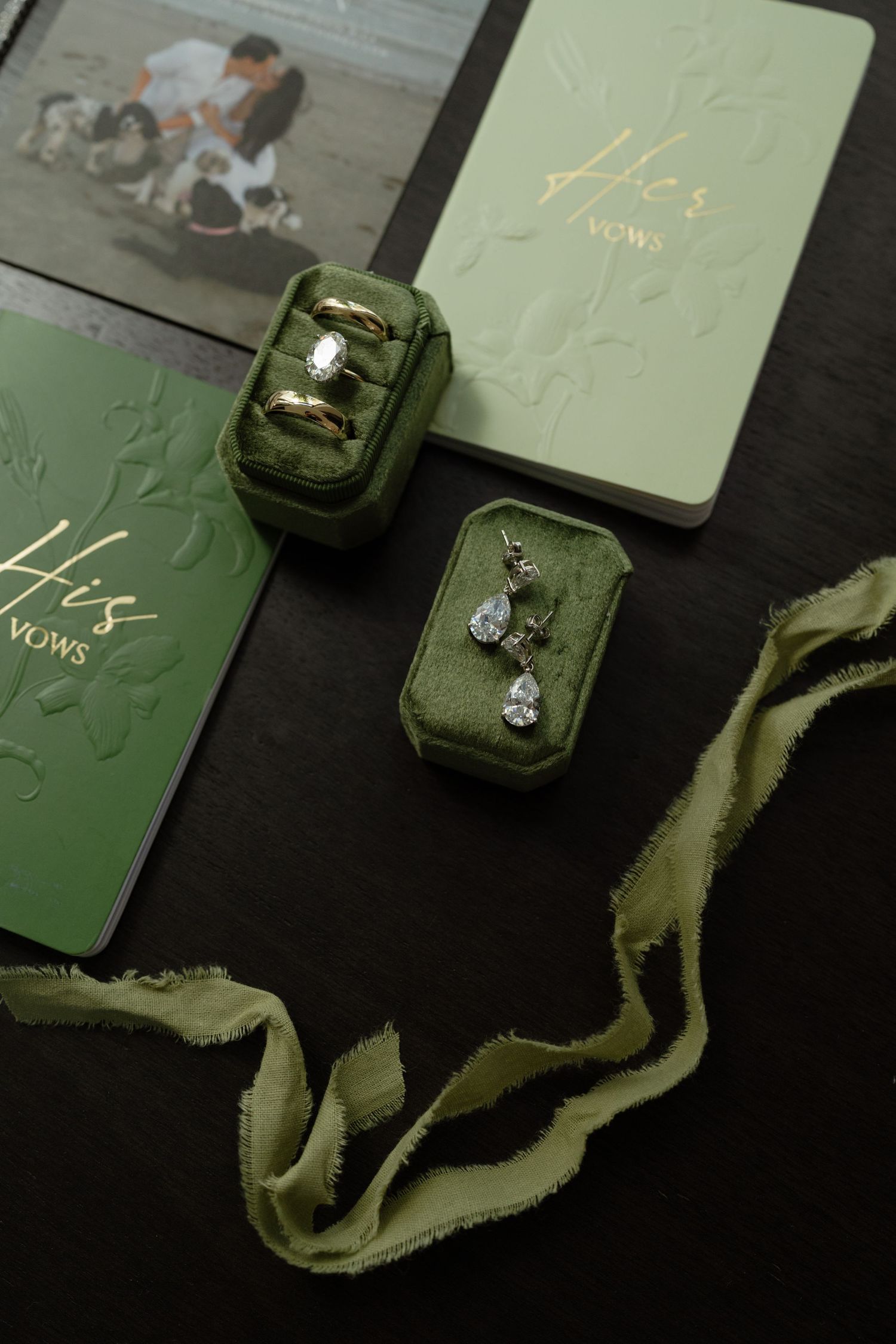 Sage green wedding stationery and ring boxes arranged on dark background with silk ribbons.
