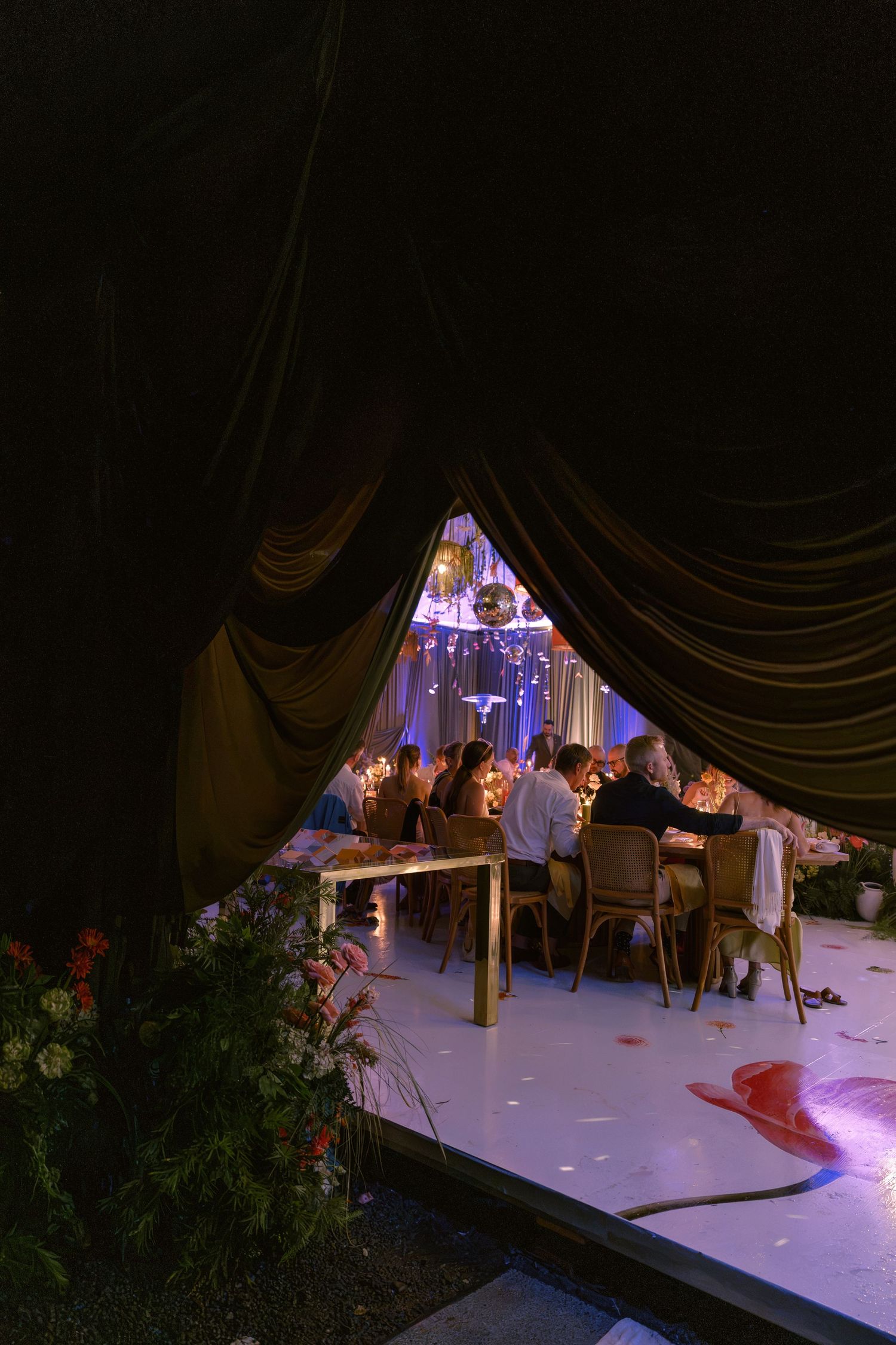 Elegant wedding reception viewed through draped curtains with guests dining by candlelight under decorative chandeliers.