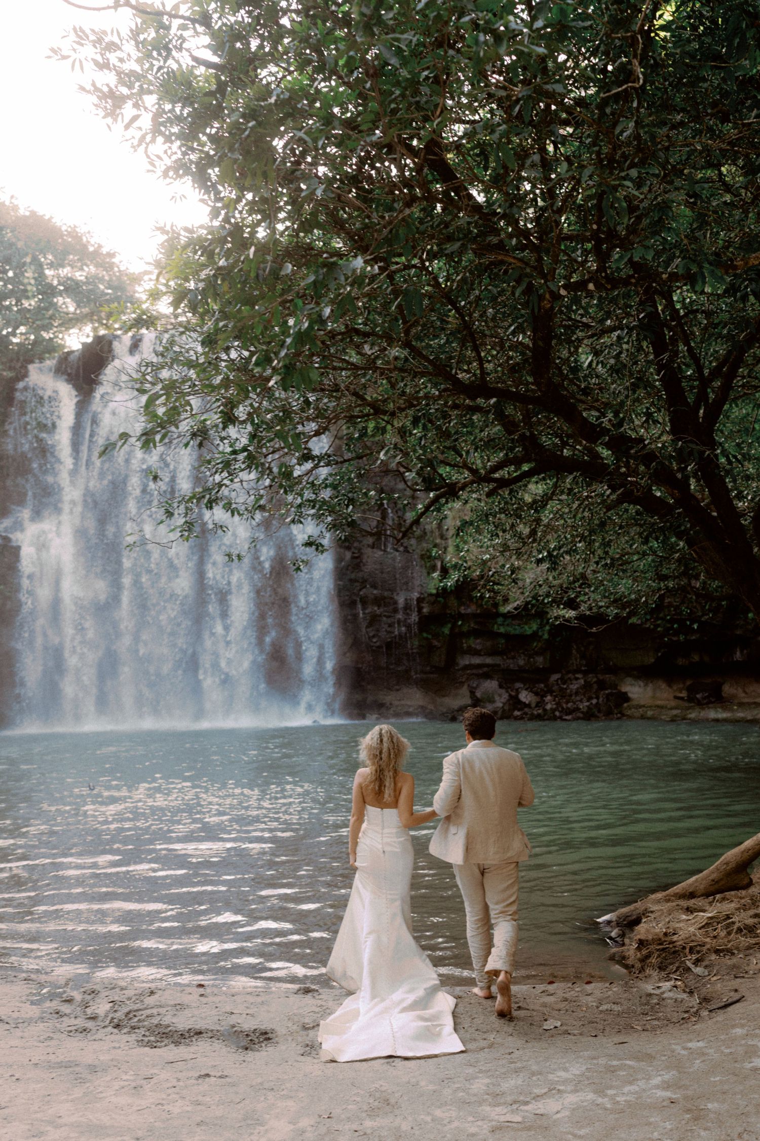 A couple in white stands by a serene waterfall pool surrounded by green foliage.