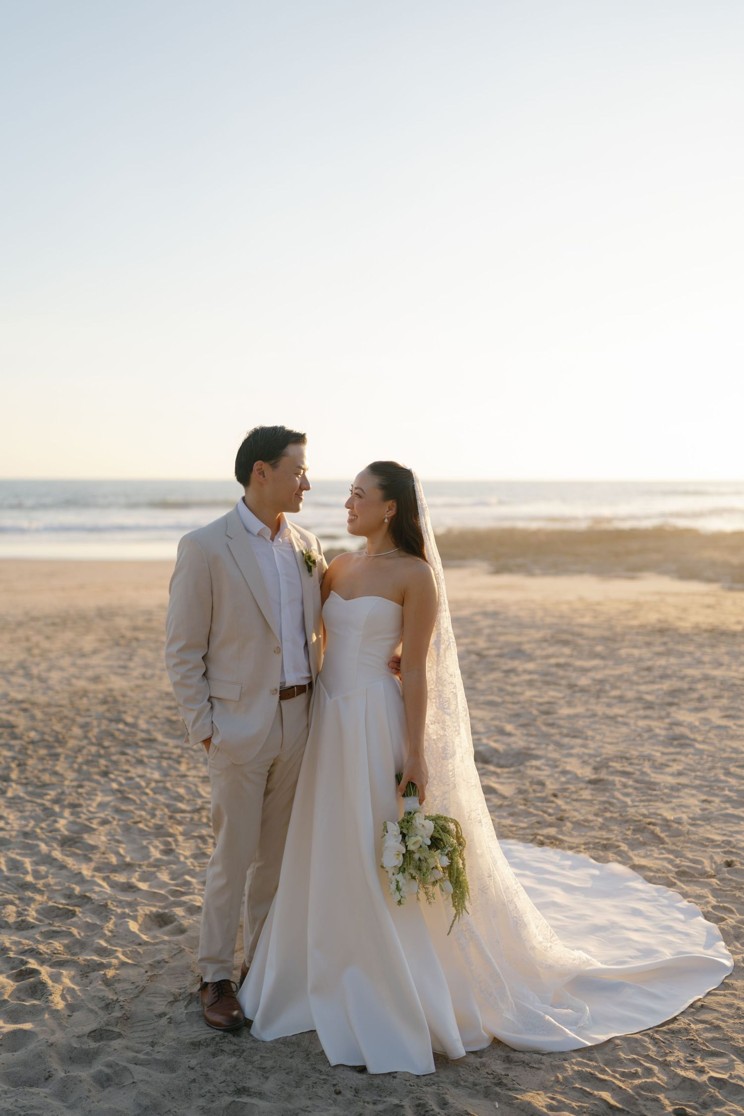A romantic beach wedding photoshoot with a couple in formal attire posing on the sand at sunset.