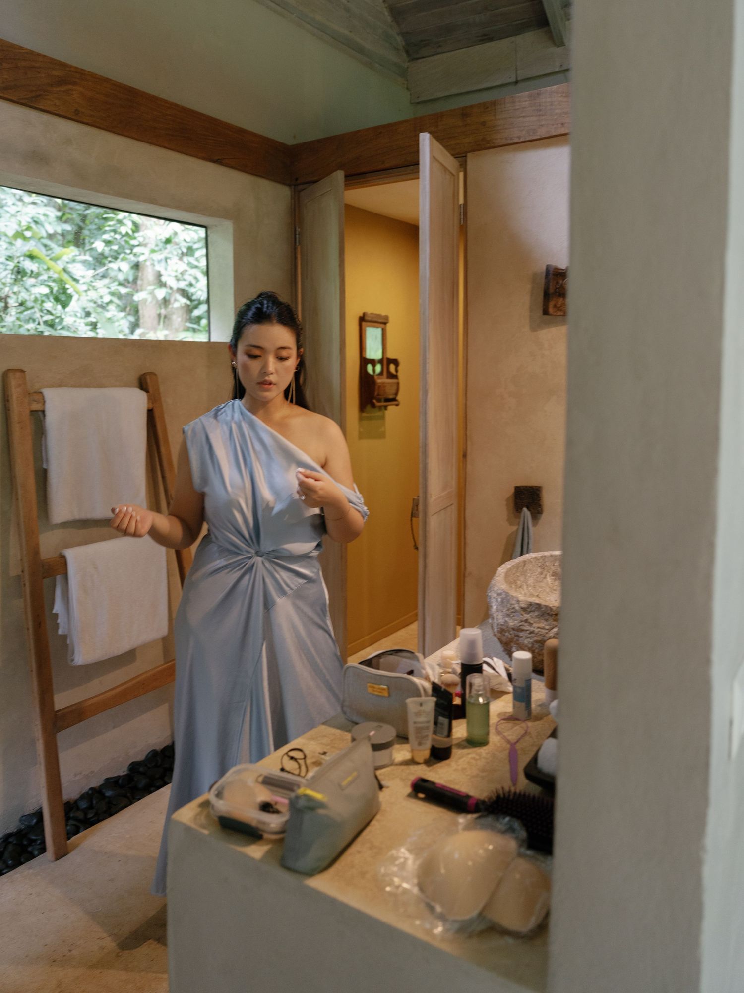 Woman in elegant blue dress standing at bathroom vanity with toiletries and mirror.