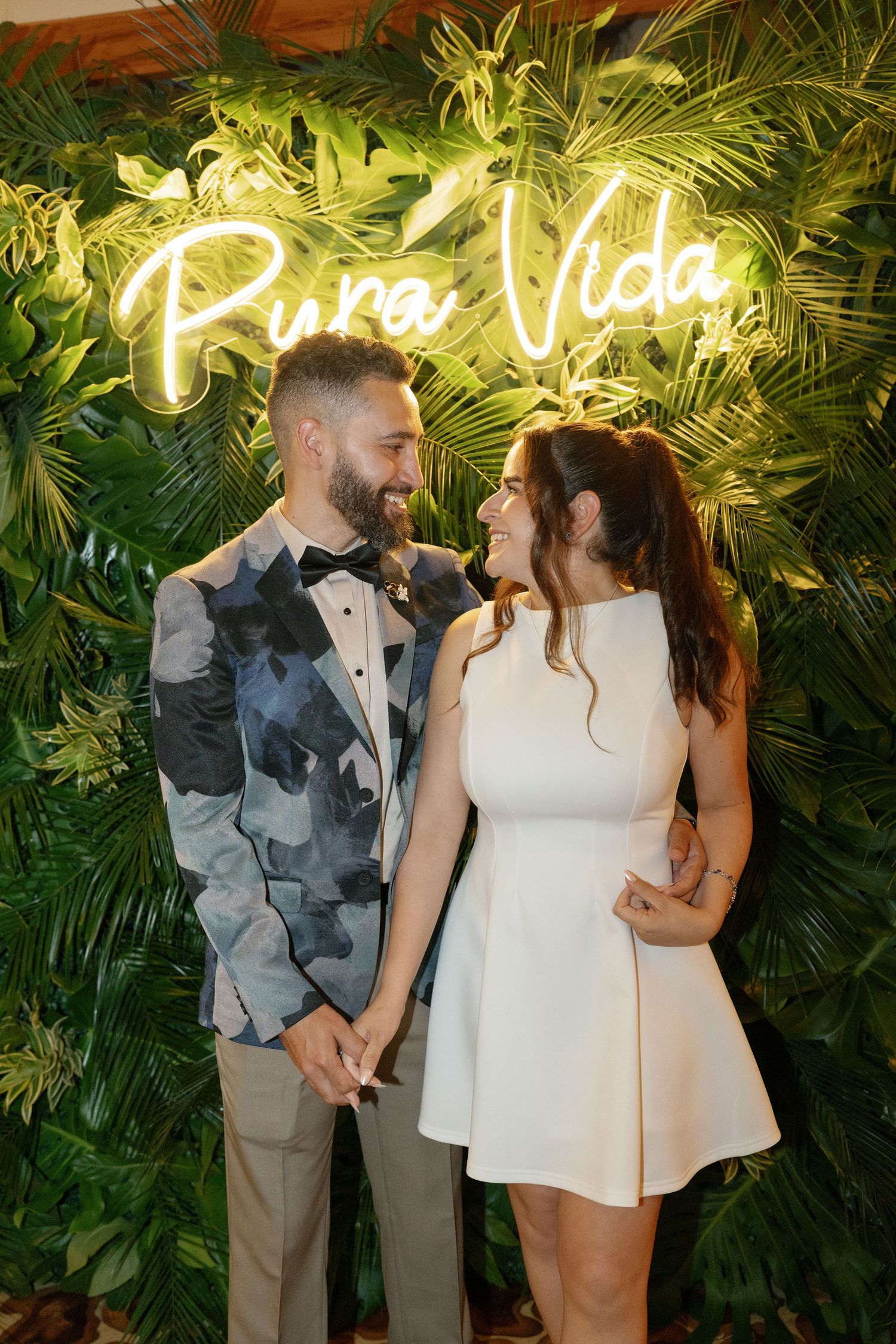 Couple poses in front of a tropical leaf wall with neon 'Pura Vida' sign.