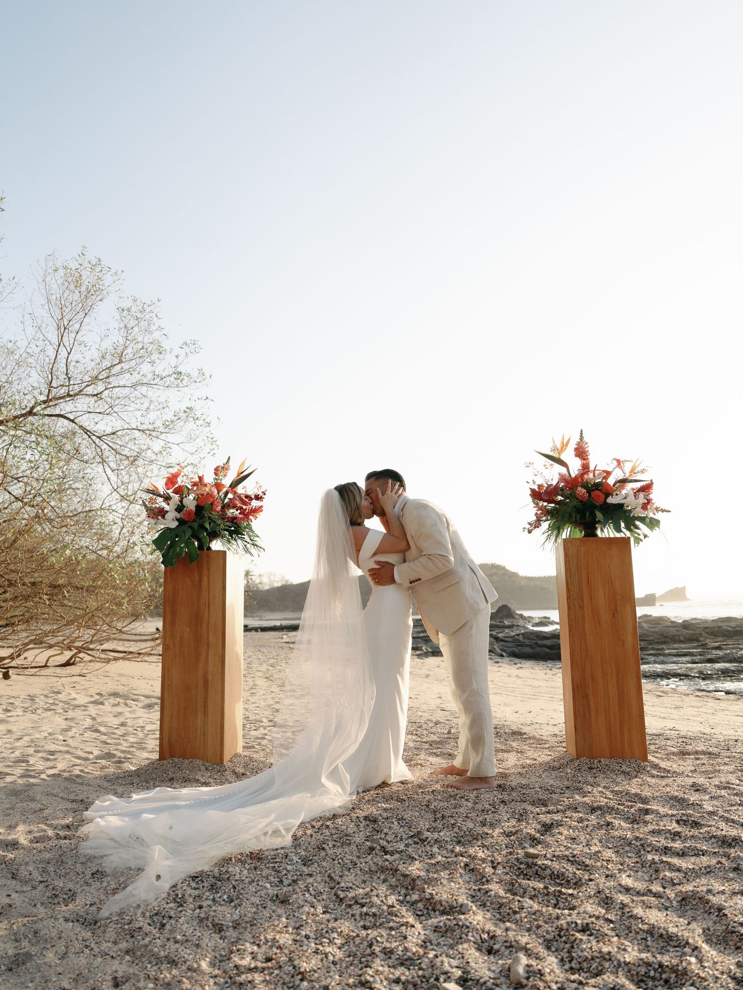 A romantic beach wedding ceremony setup with two wooden pillars adorned with pink floral arrangements during golden hour.