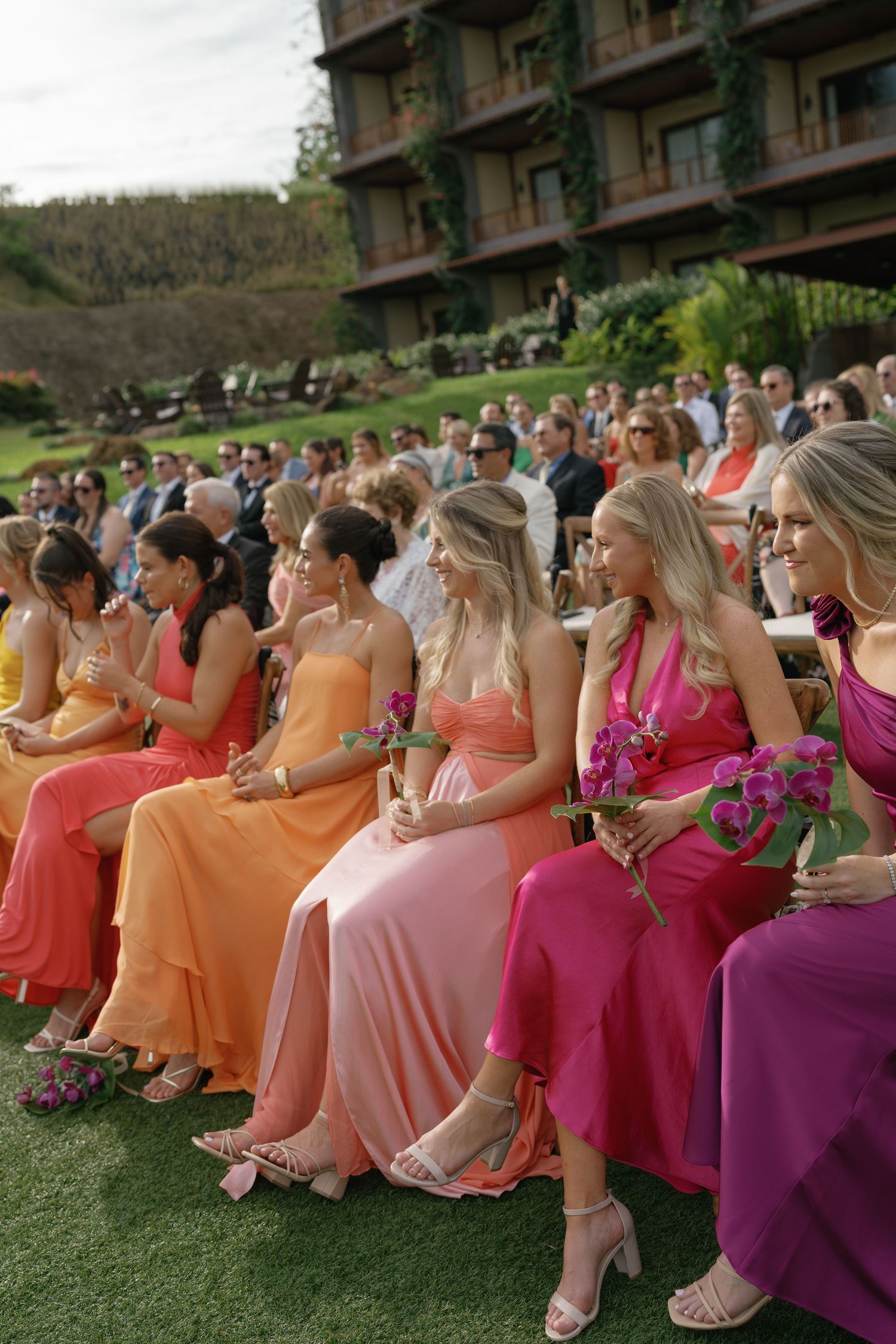 Bridesmaids in vibrant coral, pink and purple dresses seated in rows at an outdoor venue.