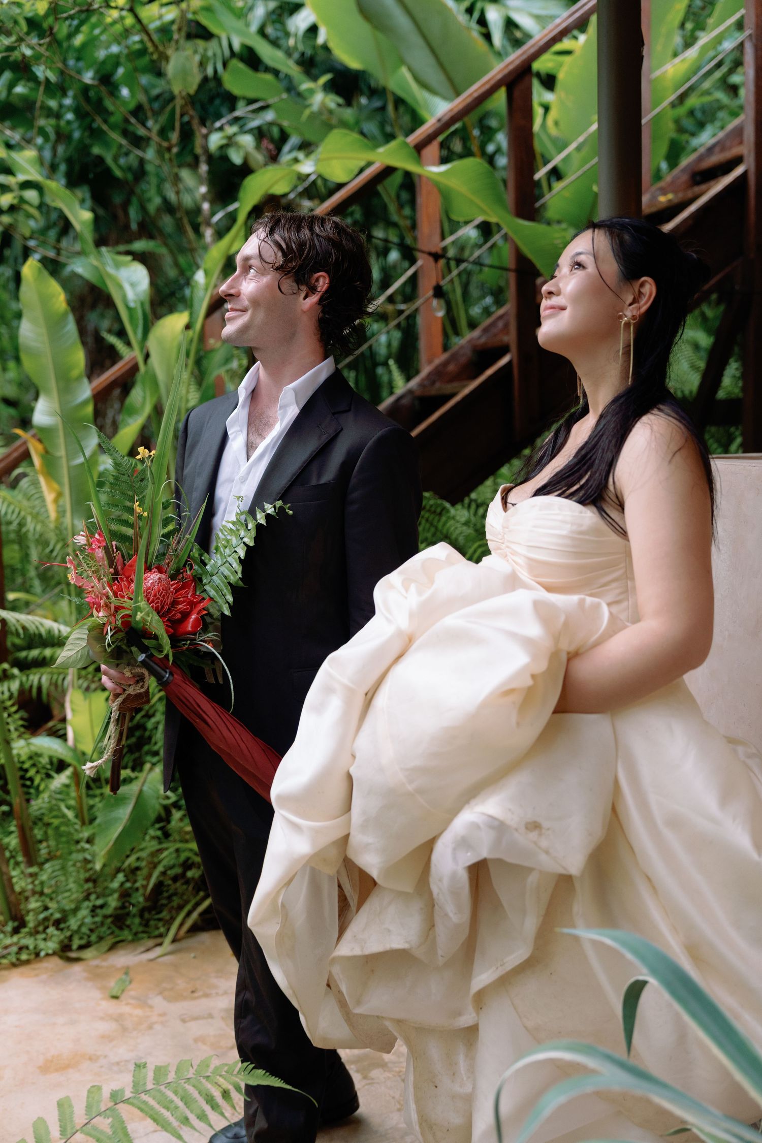 A wedding couple shares a candid moment on stairs surrounded by tropical plants.