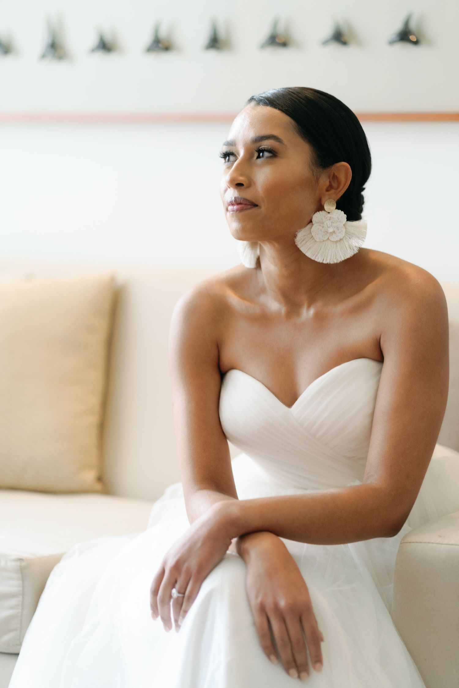 Elegant bride in white strapless wedding gown sitting on cream couch with delicate floral earrings.