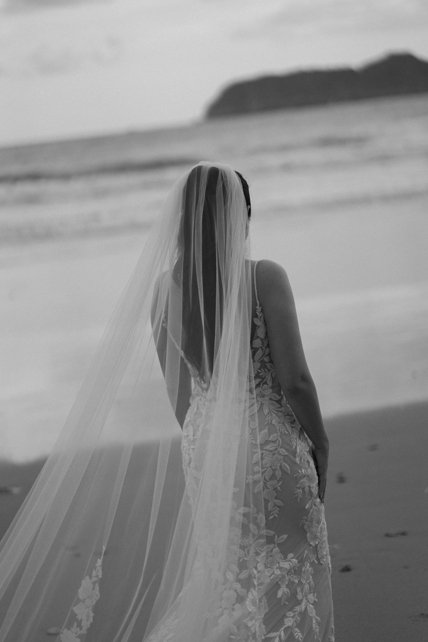 Black and white beach wedding photo featuring flowing veil in ocean breeze.
