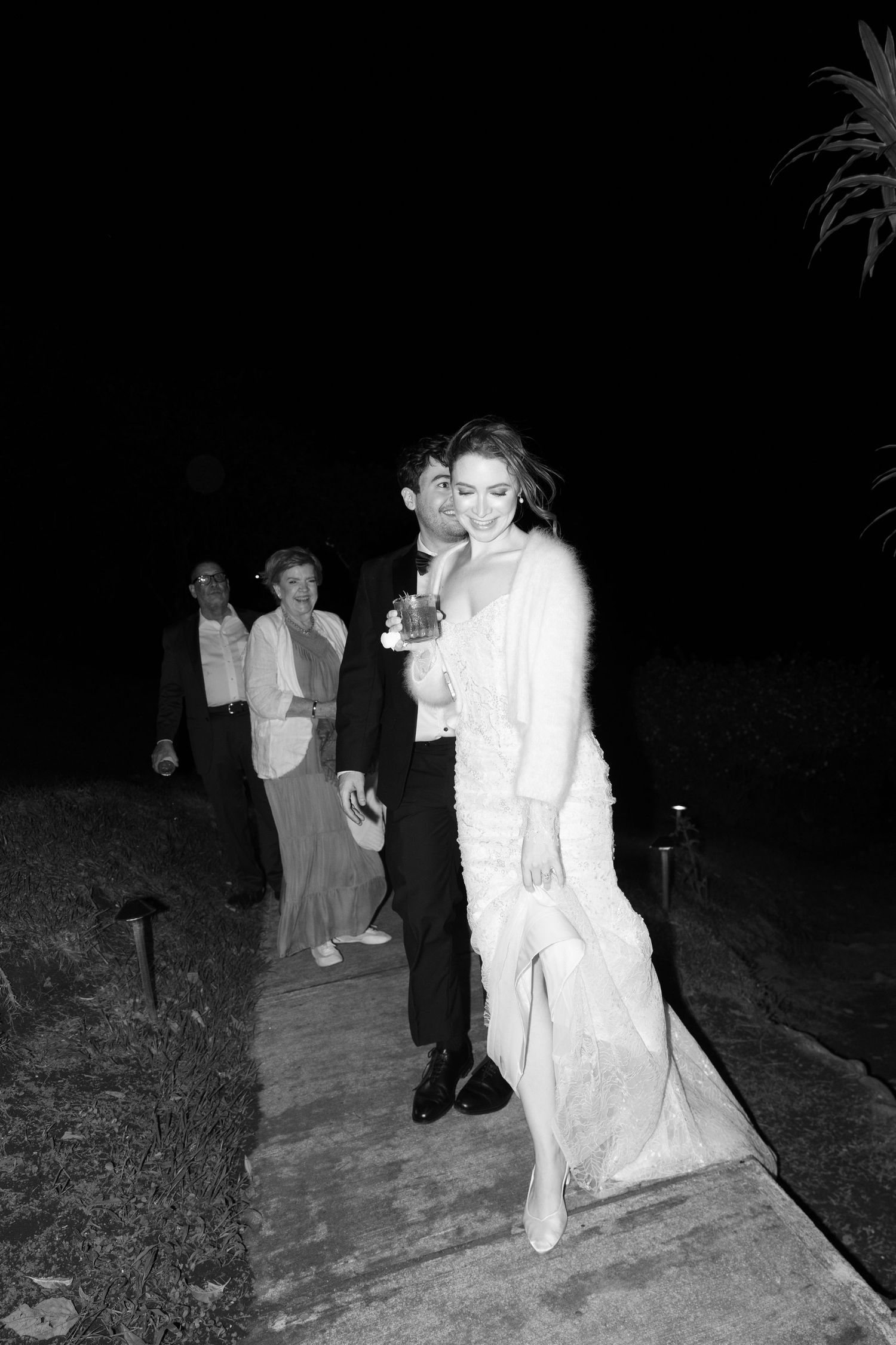 Black and white photo of a bride and groom walking at night, with wedding guests following behind them.